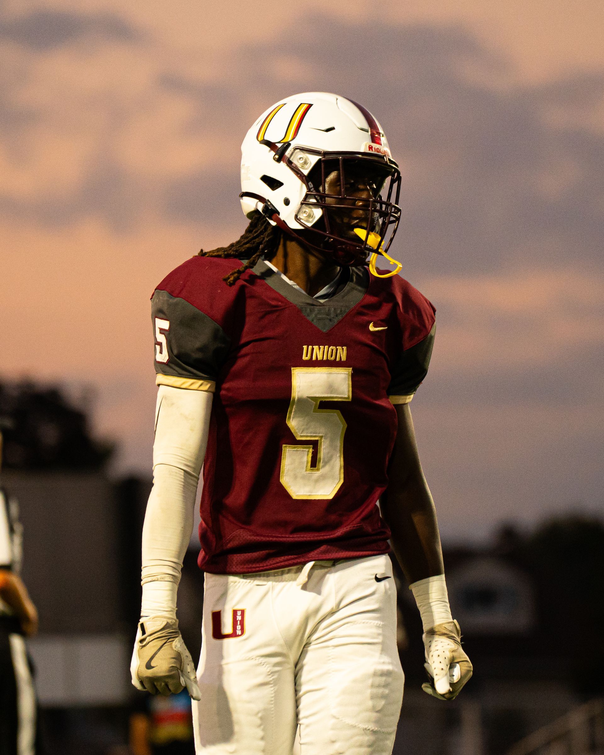 Football player in maroon jersey number 5 and white helmet standing on the field at sunset