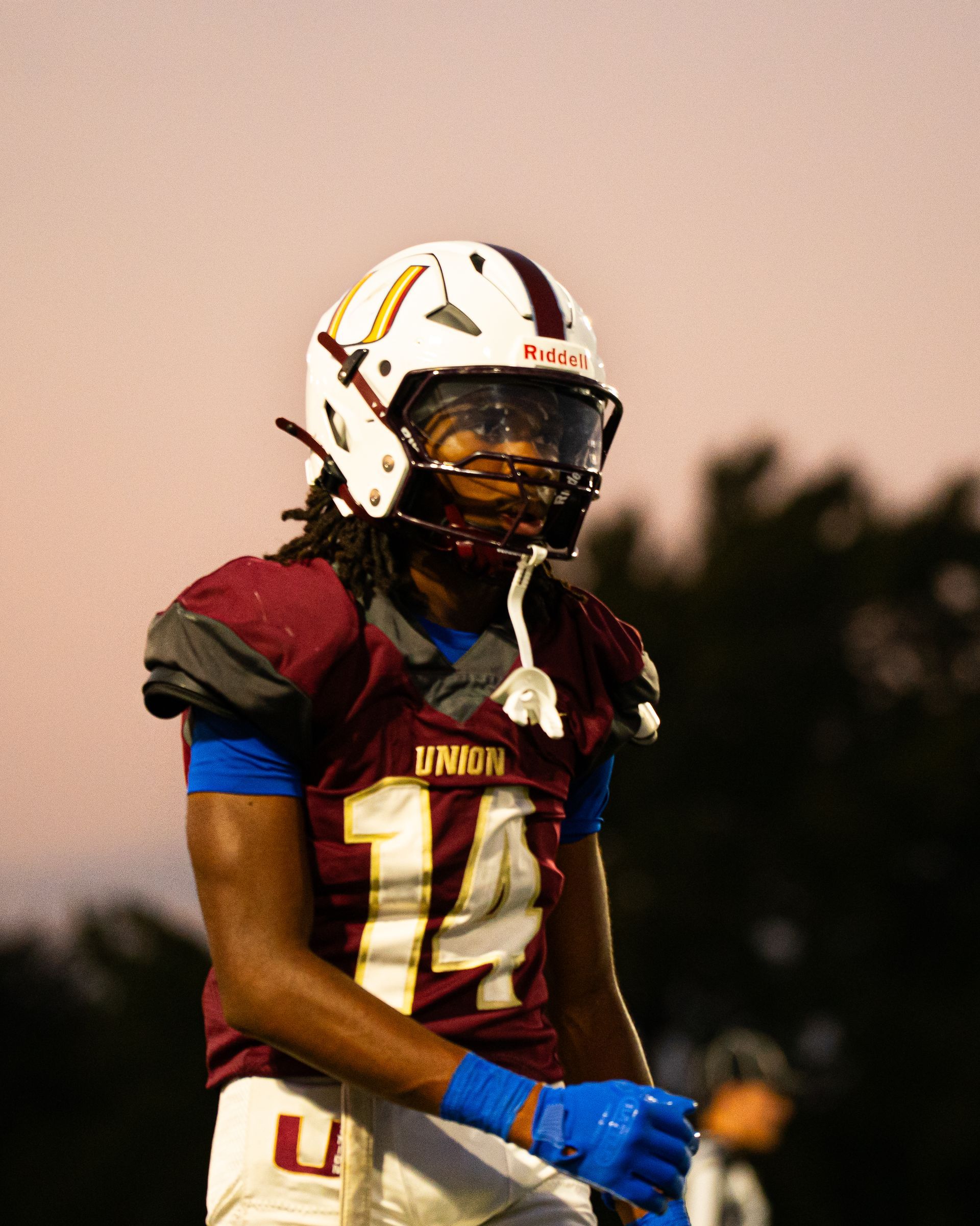 Football player in maroon and gold uniform and white helmet standing on field at dusk