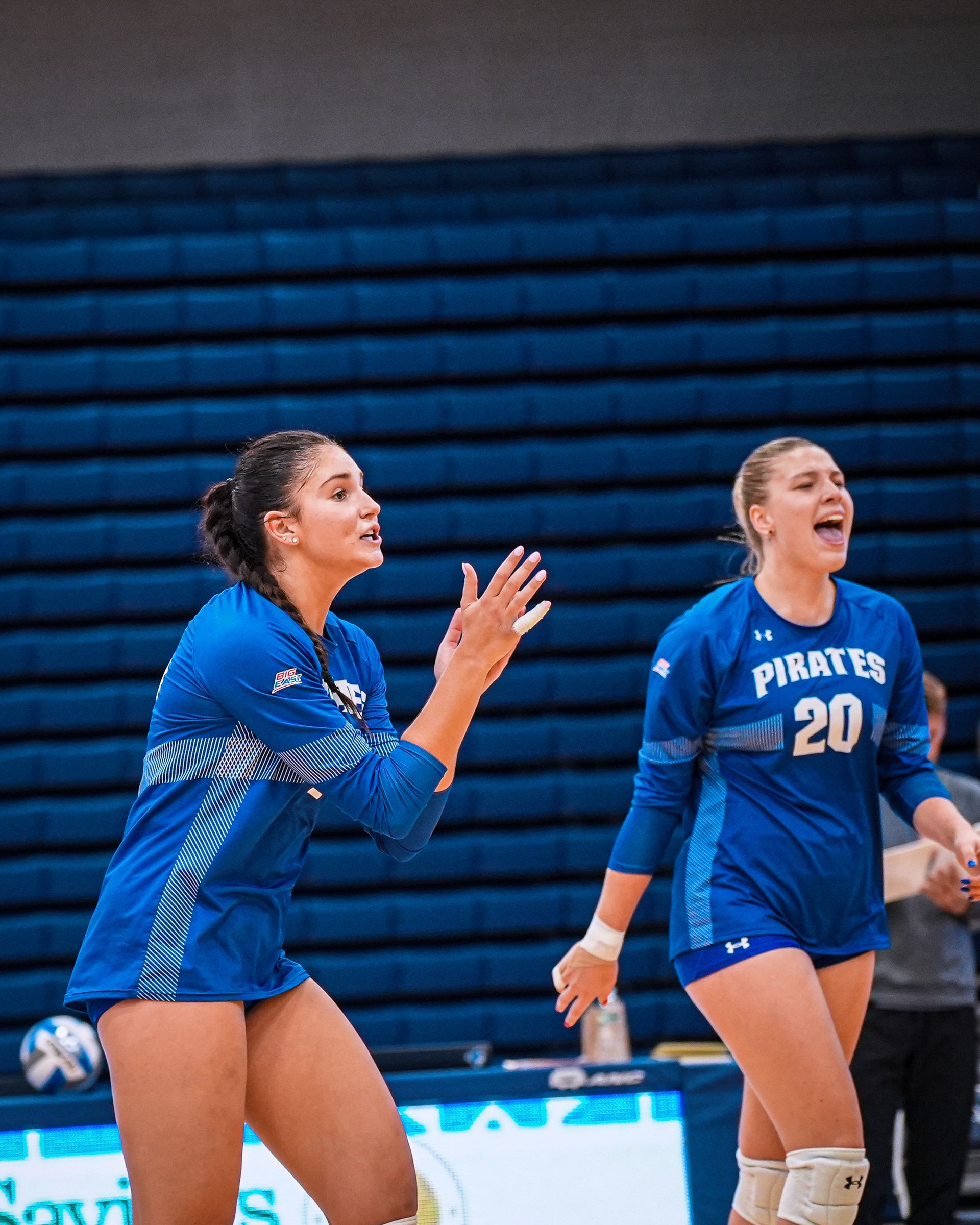 Two blue-clad volleyball players cheering on the court in front of blue bleachers