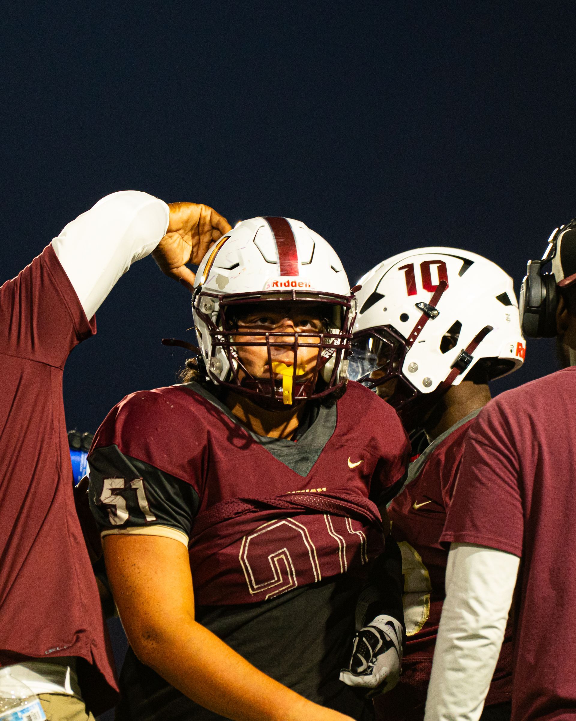 Football player in maroon uniform and white helmet standing with teammates on the sideline