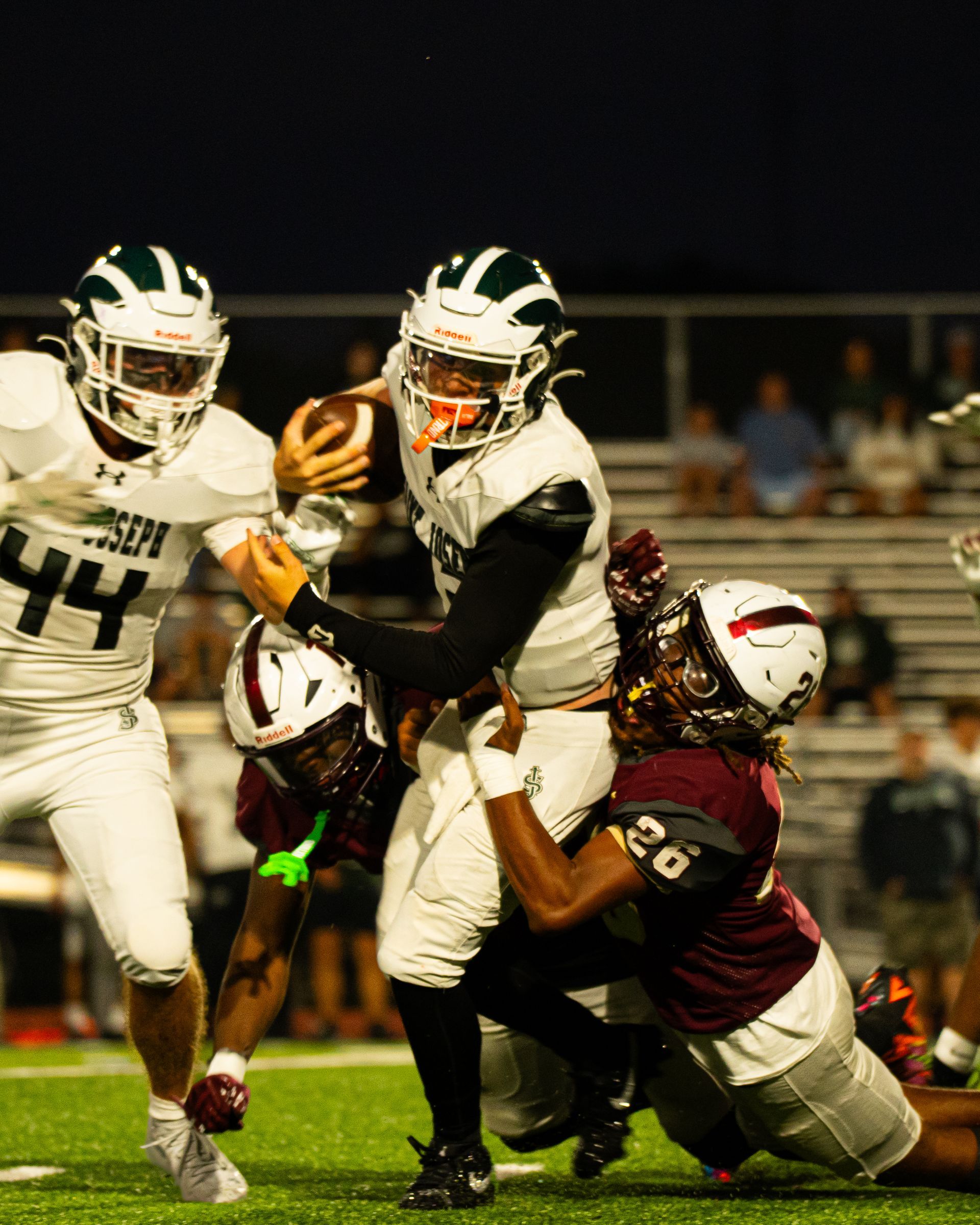 High school football players in white and maroon collide as one runs with the ball under stadium lights.
