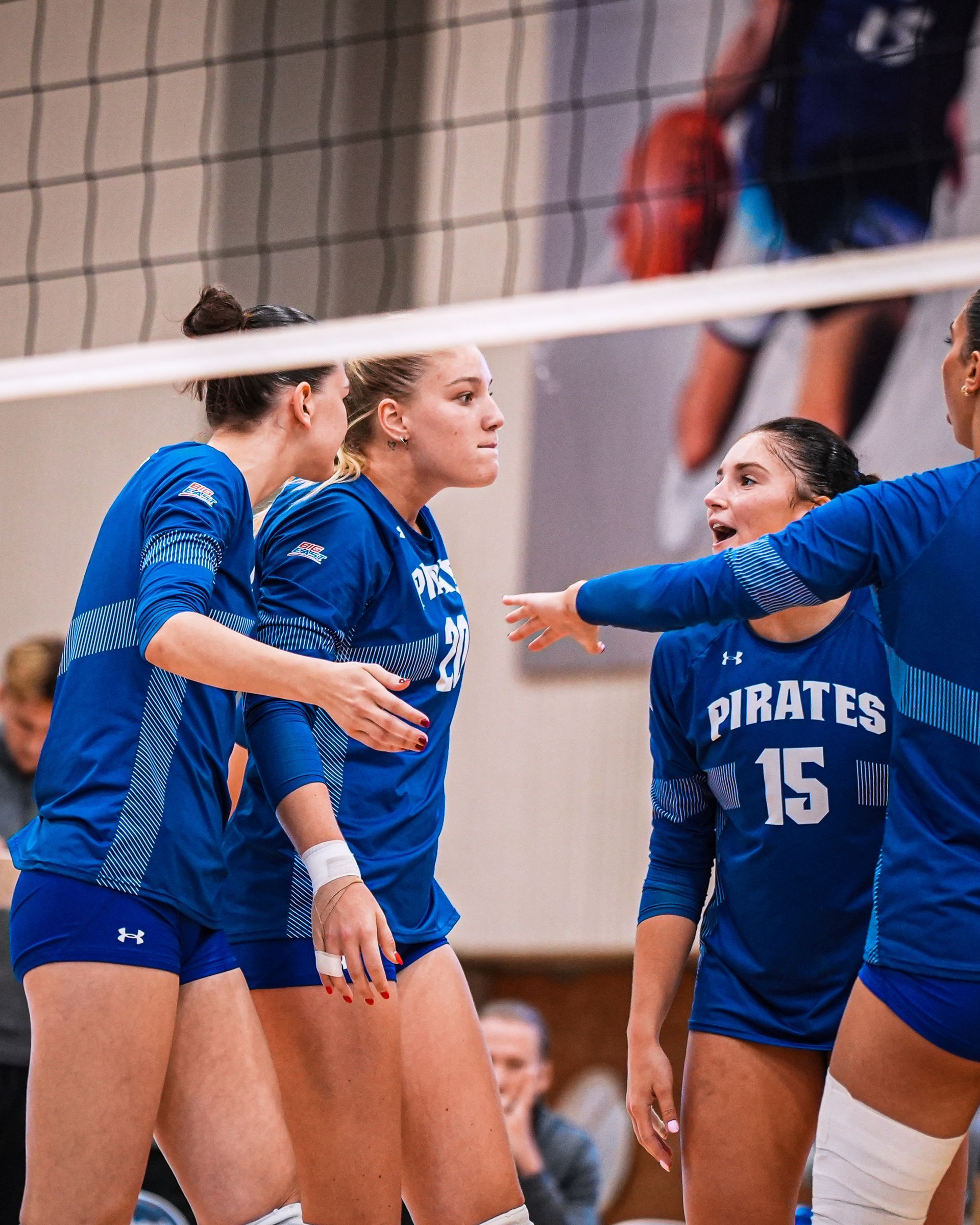 Blue-clad volleyball players huddle on court during a match, with net in foreground