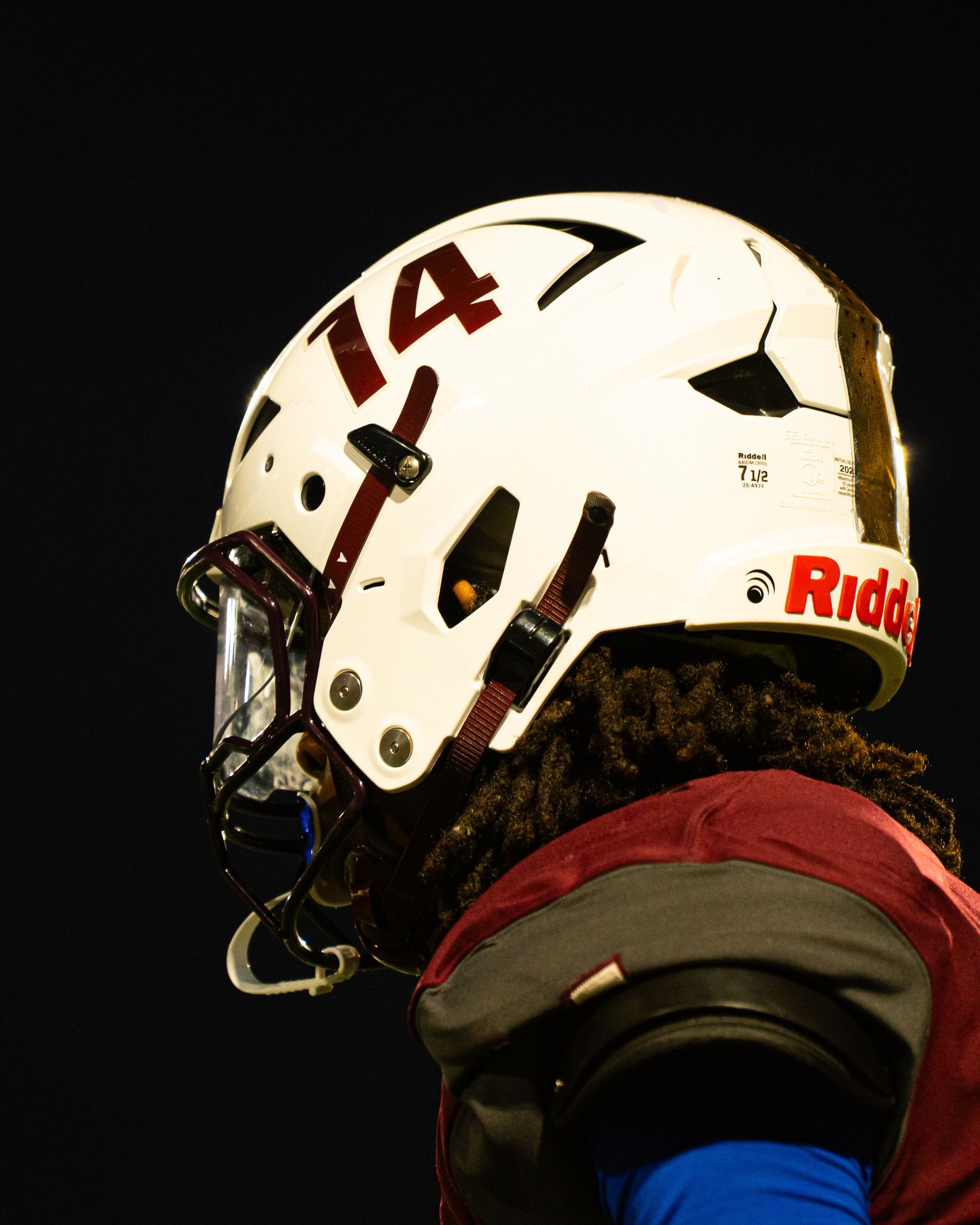 Football player in white helmet with maroon number 14, seen in profile against a dark background