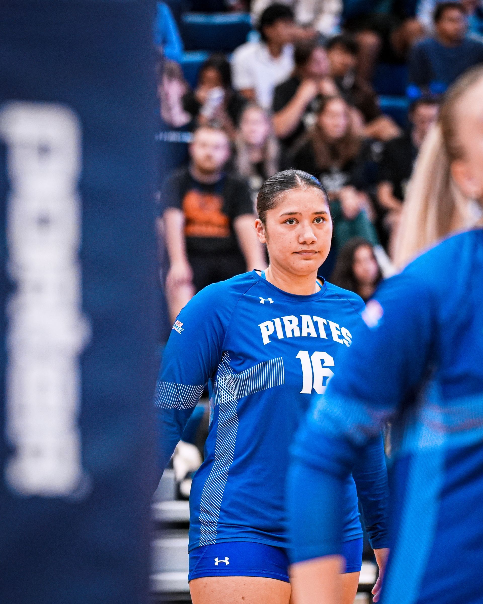 Volleyball player in blue Pirates uniform stands on court, with blurred spectators in the background