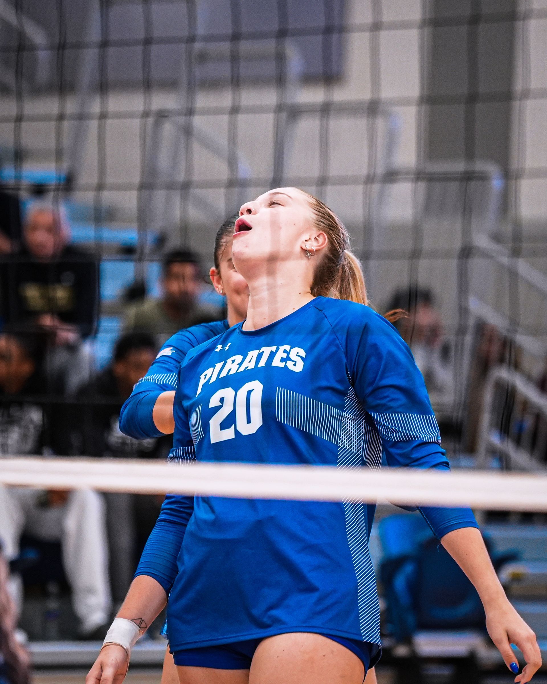 Volleyball player in blue jersey, number 20, looking upward during a game behind the net
