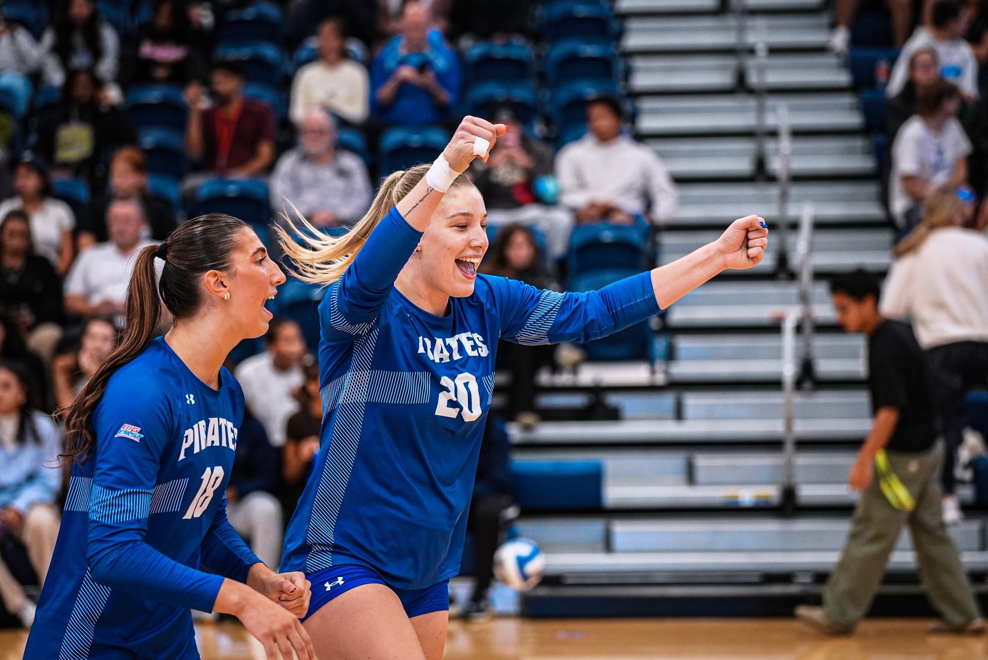 Two volleyball players in blue jerseys celebrating on the court, with spectators in the bleachers behind them