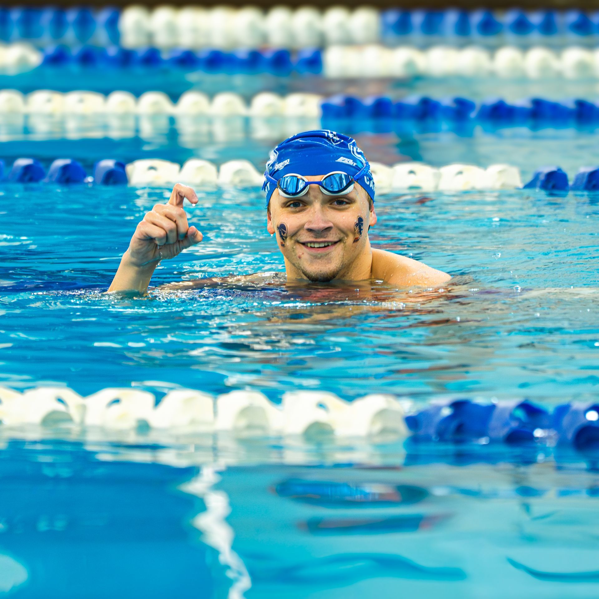 Swimmer in blue cap and goggles gives a thumbs-up in a pool with lane dividers.