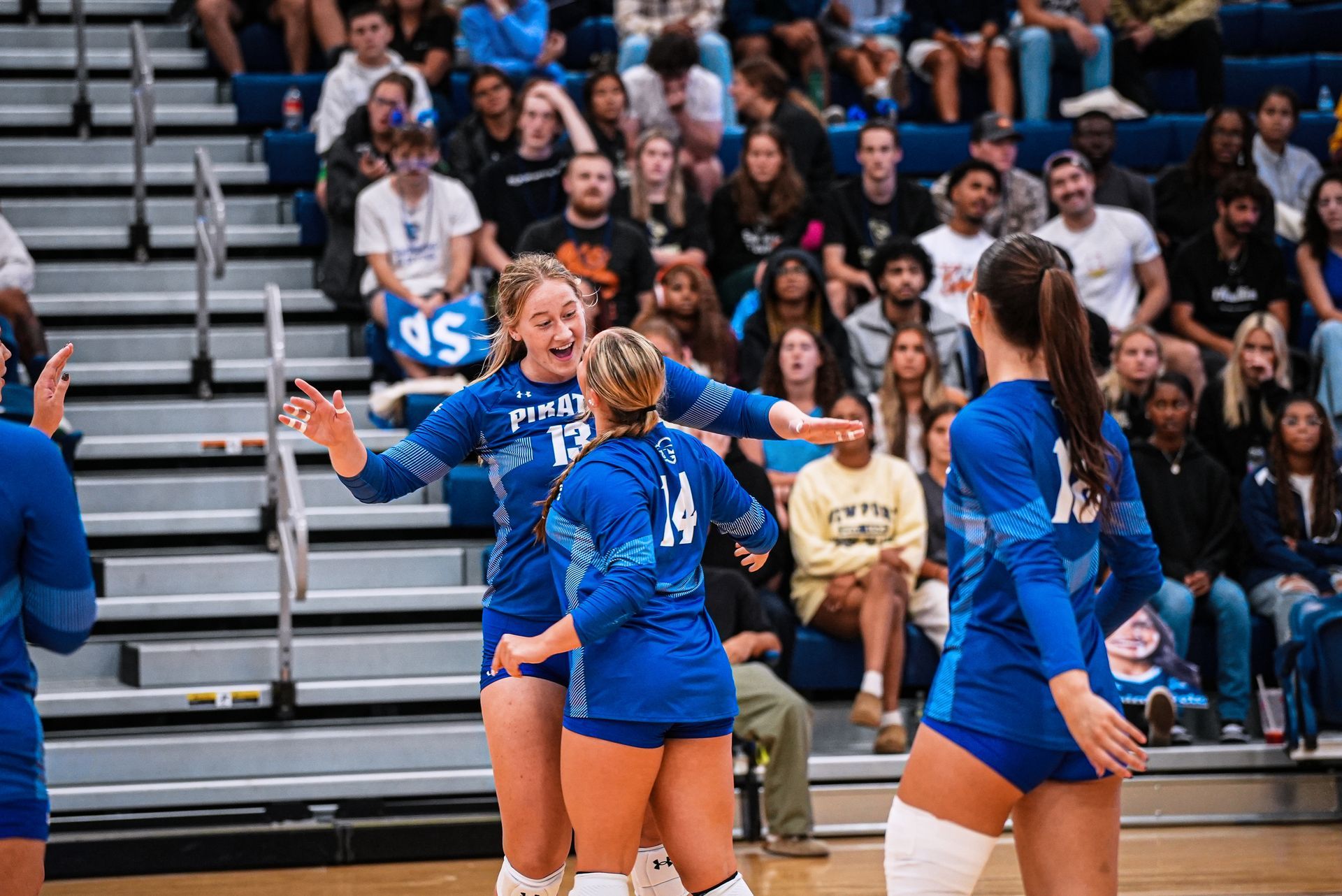 Volleyball players in blue uniforms huddle on the court with spectators in the bleachers behind them
