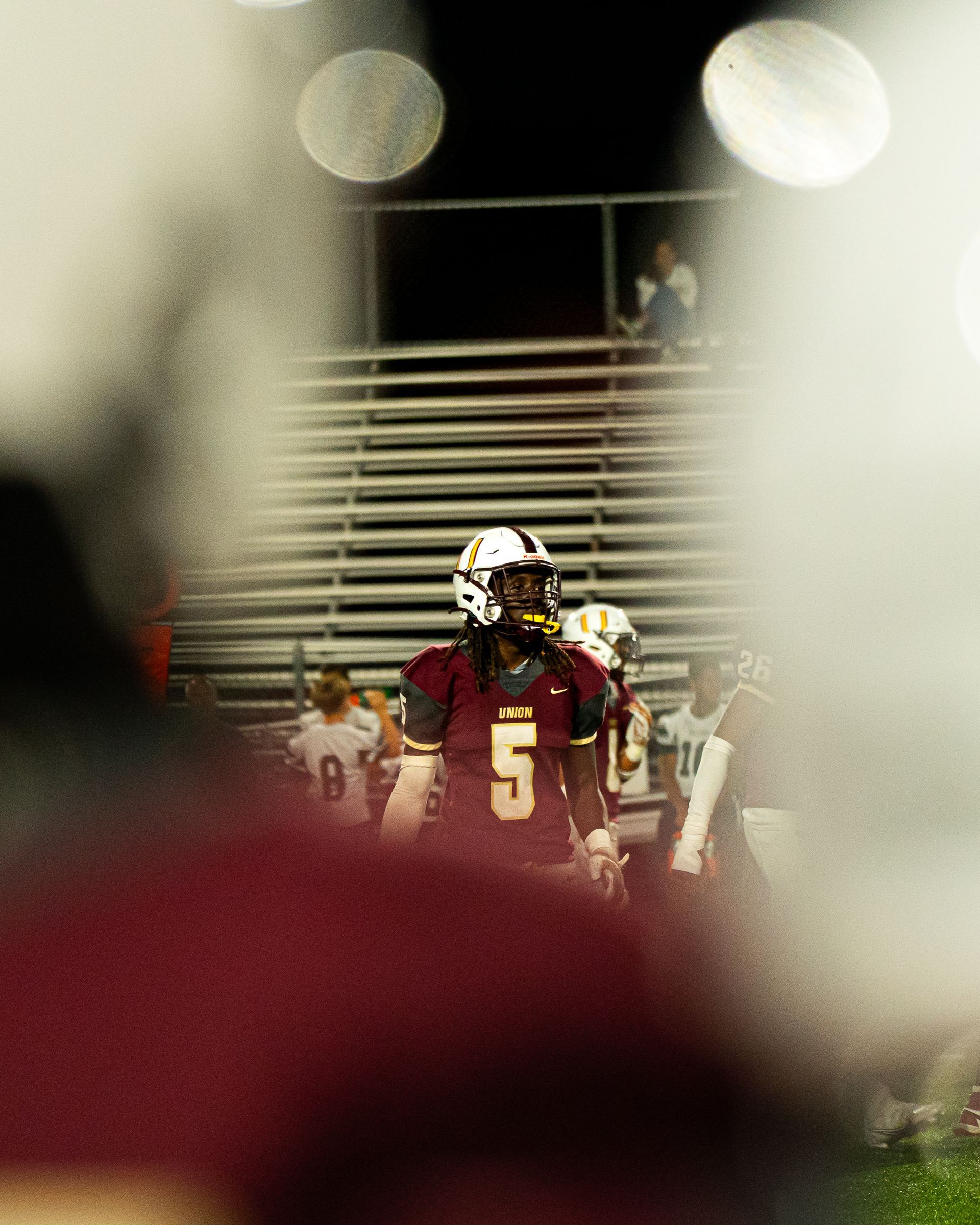 Football player in maroon jersey number 5 stands on the field near stadium steps, viewed through blurred foreground.