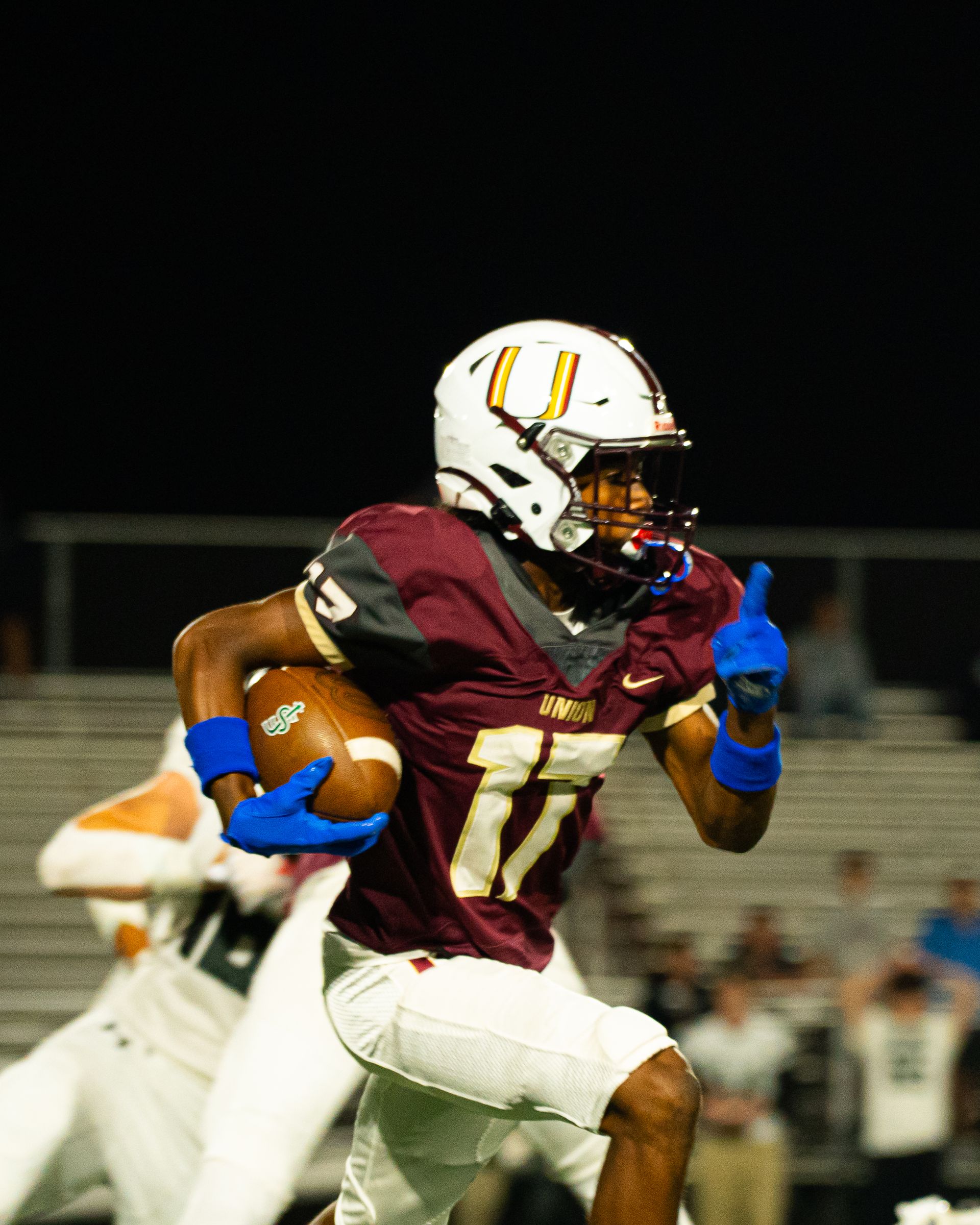 Football player in maroon jersey running with the ball under stadium lights