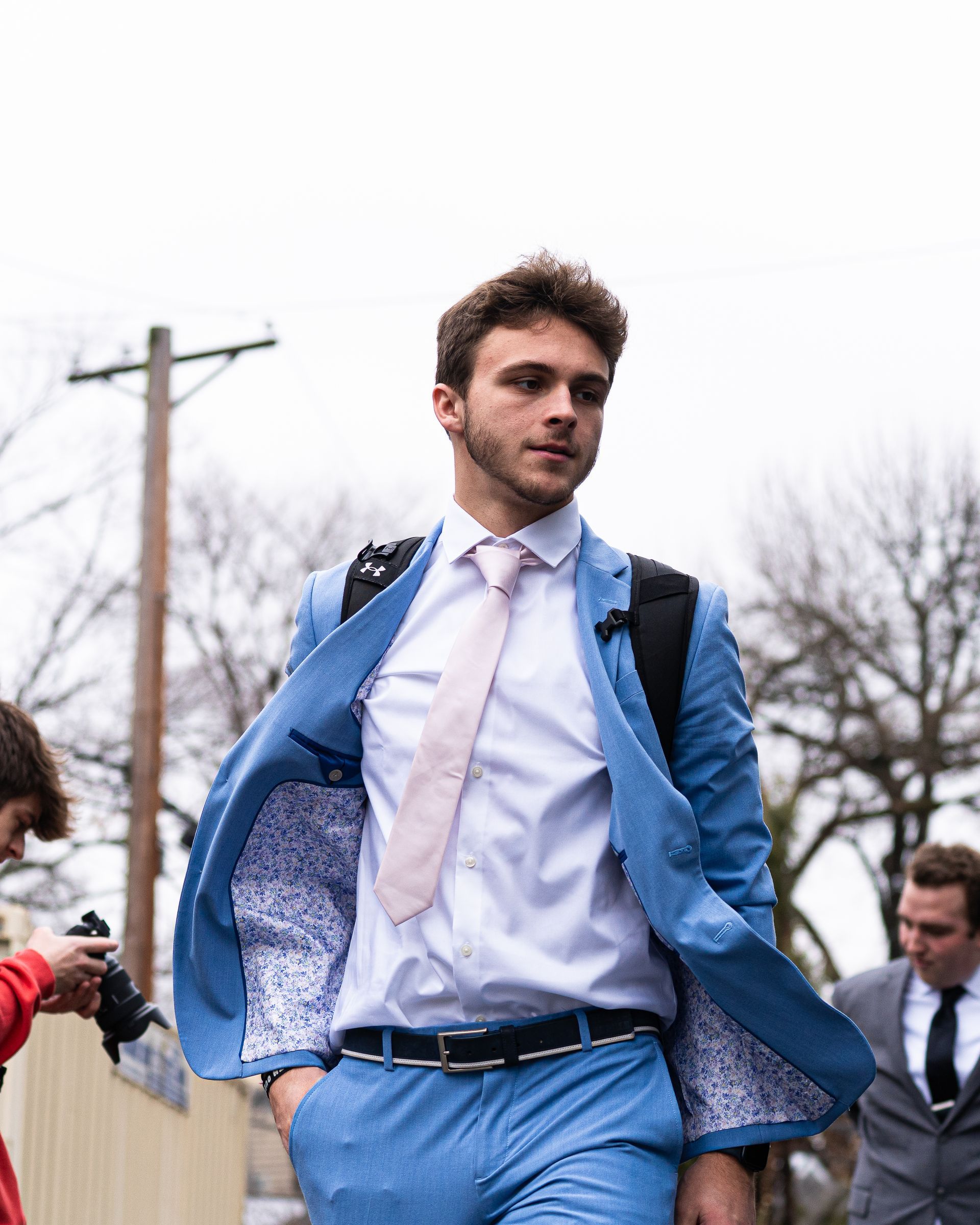 Man in a bright blue suit walks outdoors, jacket open, with trees and utility poles in the background.