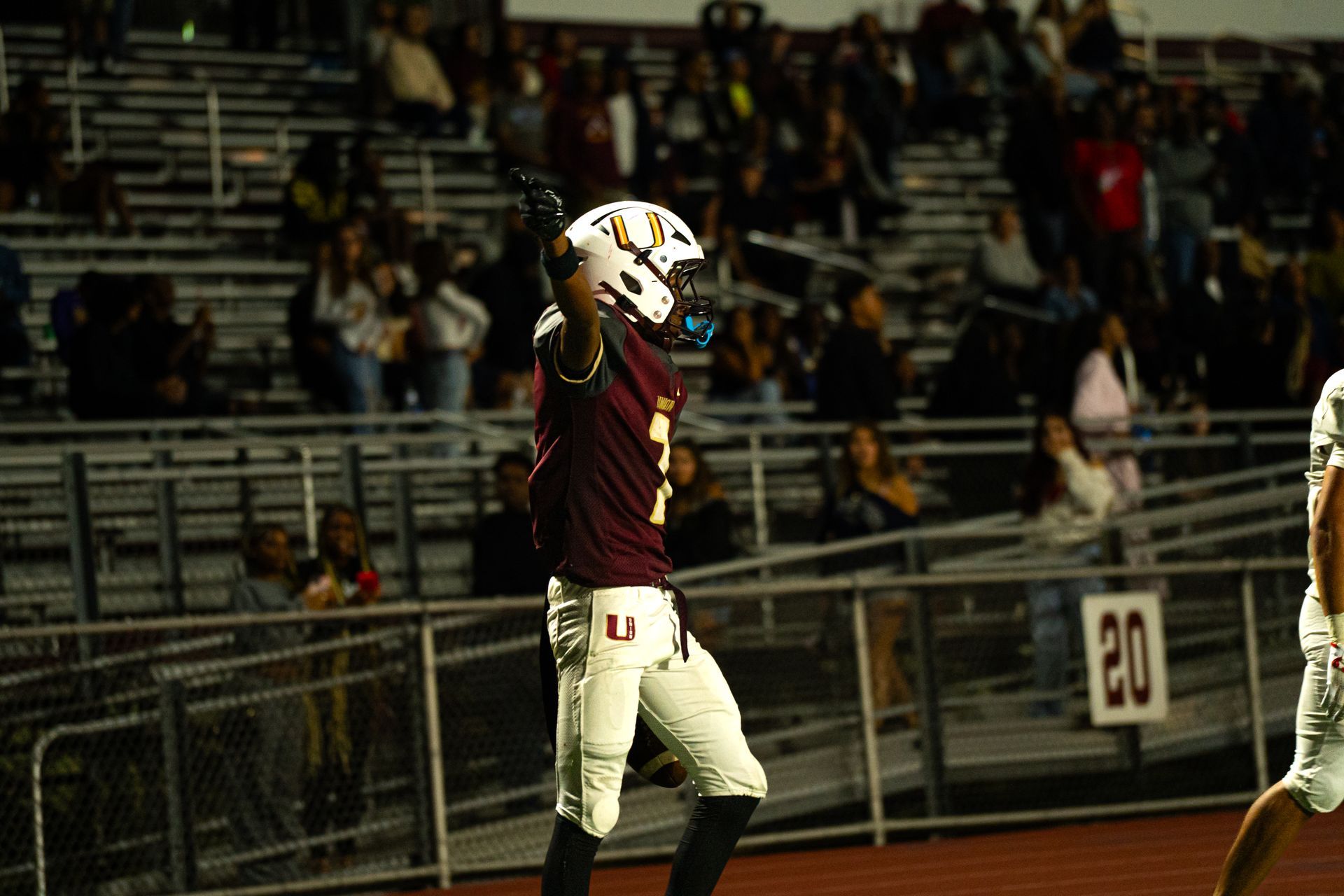 Football player in maroon jersey and white pants raises his arm on the sideline at night game
