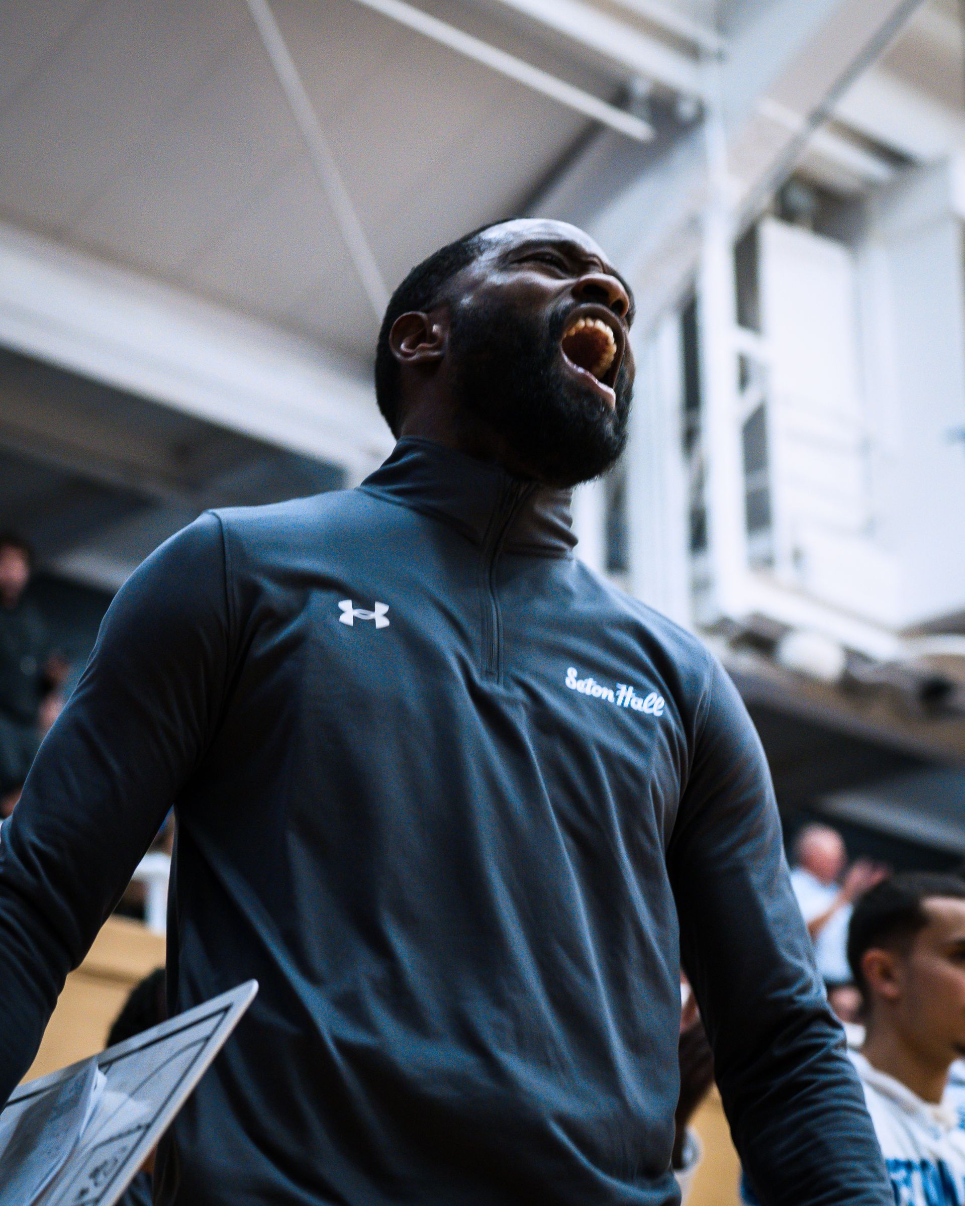 Athlete in dark Under Armour top yawns or shouts in an indoor gym, holding a clipboard.