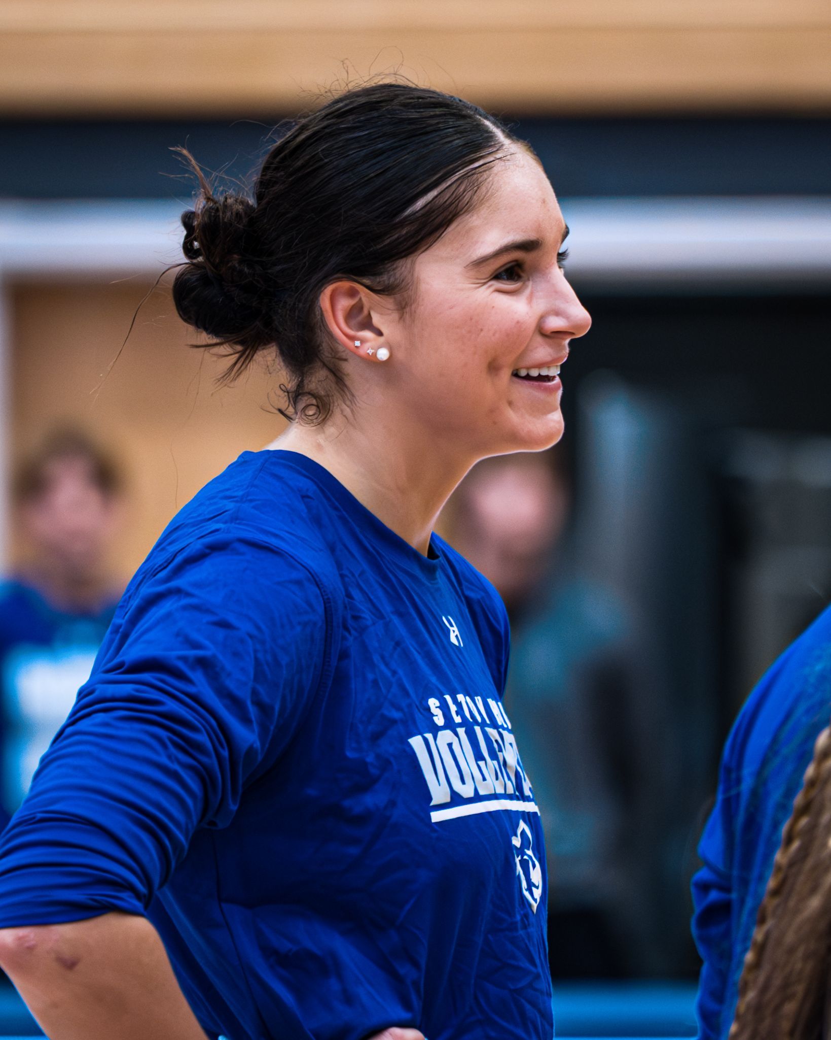 Smiling volleyball player in blue jersey standing indoors in profile, hands on hips