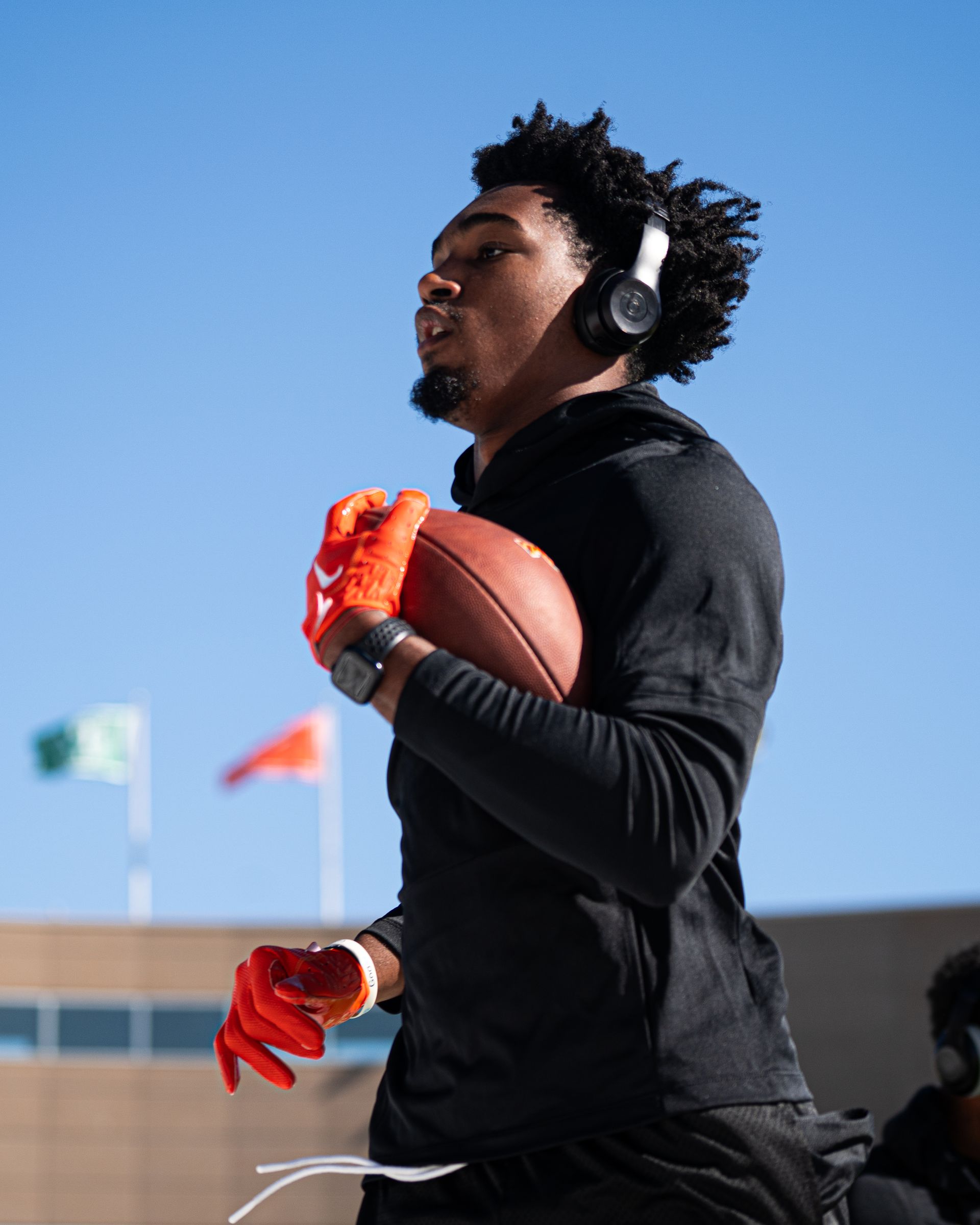 Football player in black gear holding a football, wearing orange gloves and headphones outdoors under a blue sky