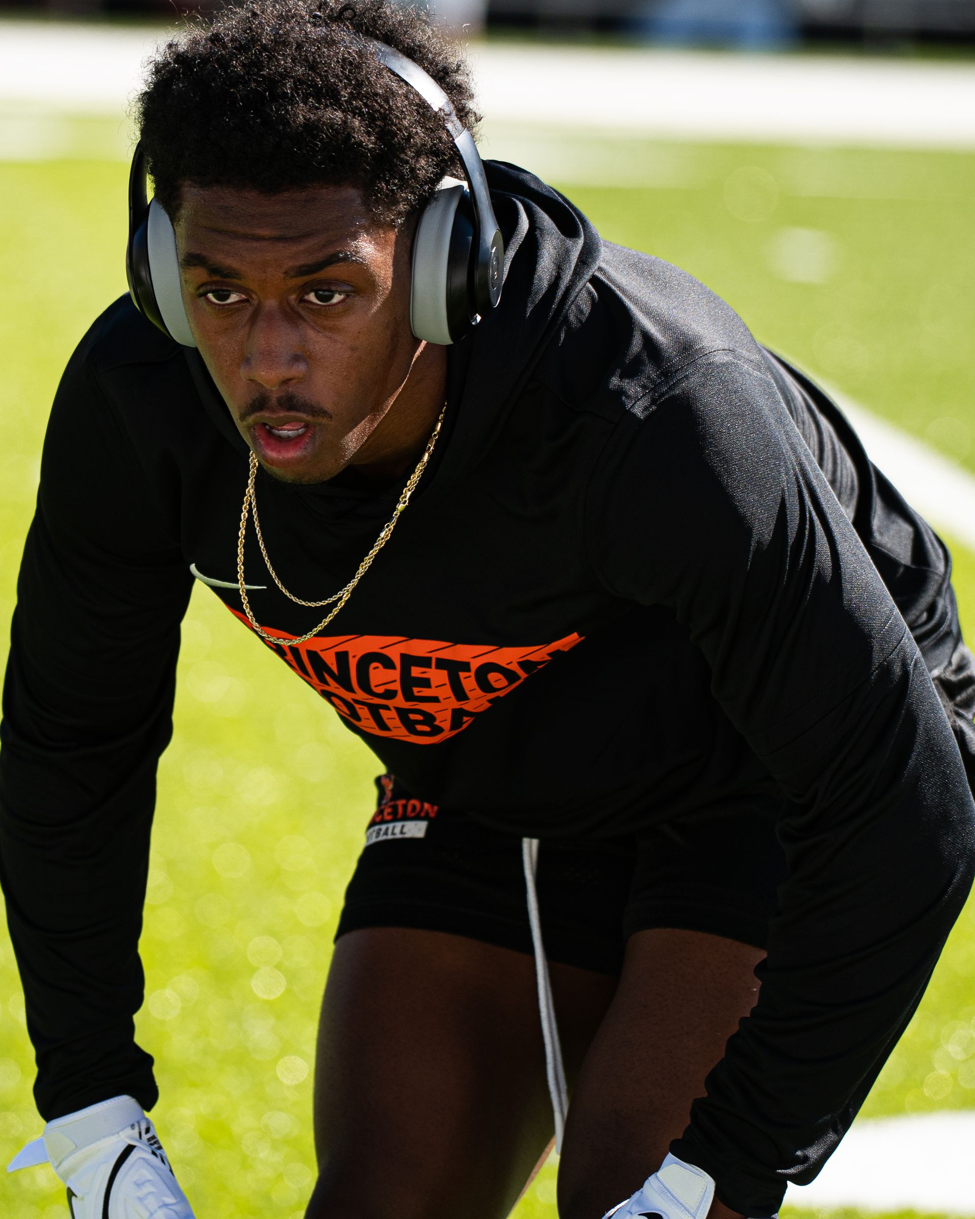 Football player in black practice gear crouches on a sunny field, wearing headphones and white gloves.