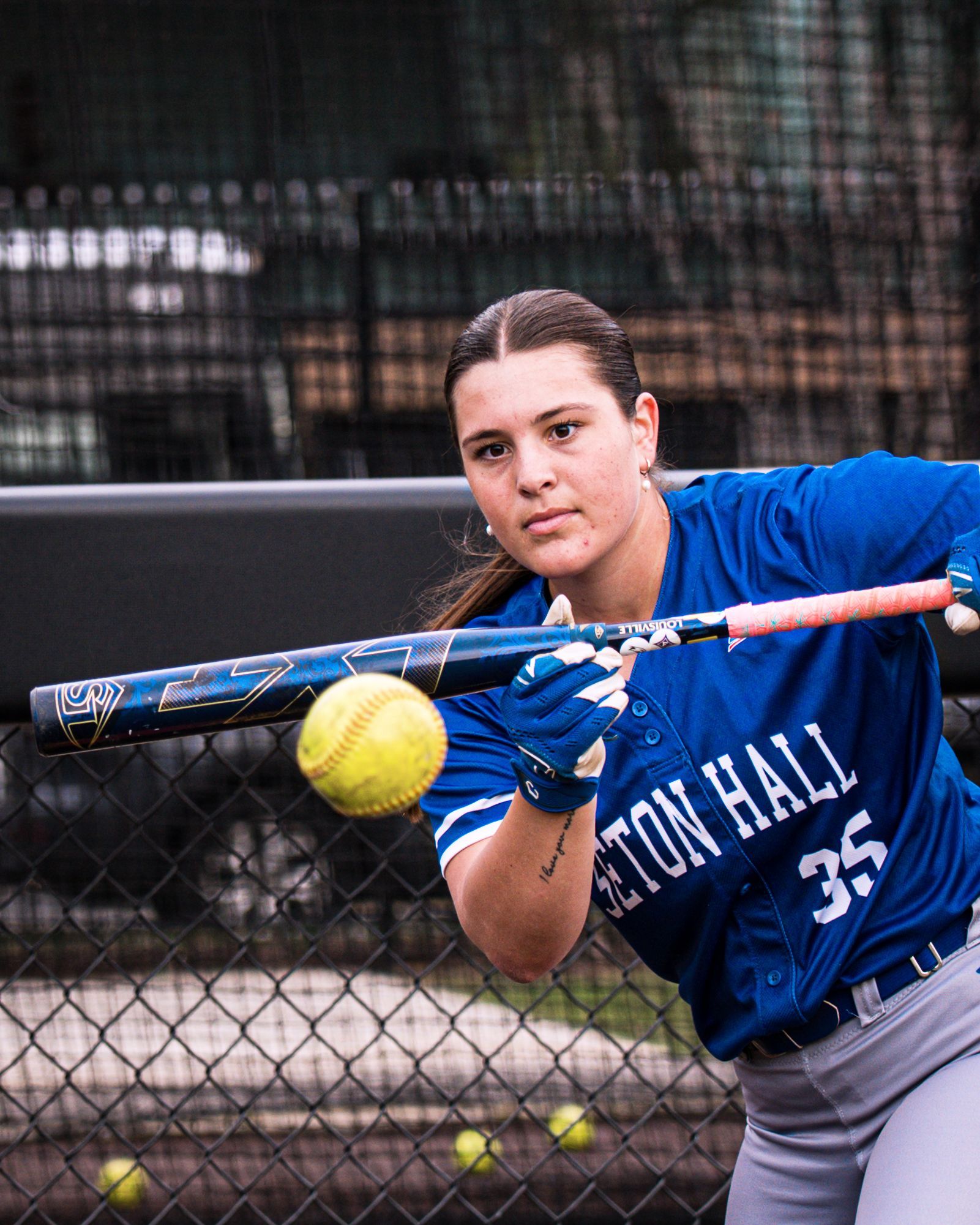 Softball player in blue swings at a yellow ball near a chain-link fence