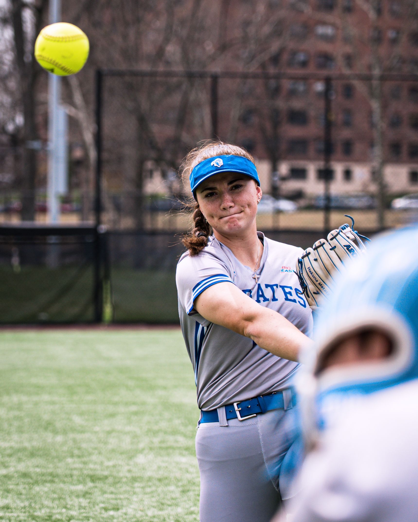 Softball player in gray and blue swings at a yellow ball on a field with a blurred teammate in foreground