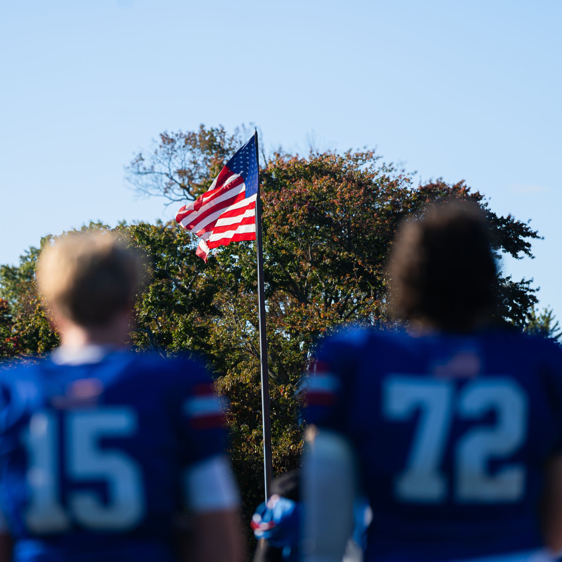 Football players in blue jerseys stand before a waving American flag on a field.