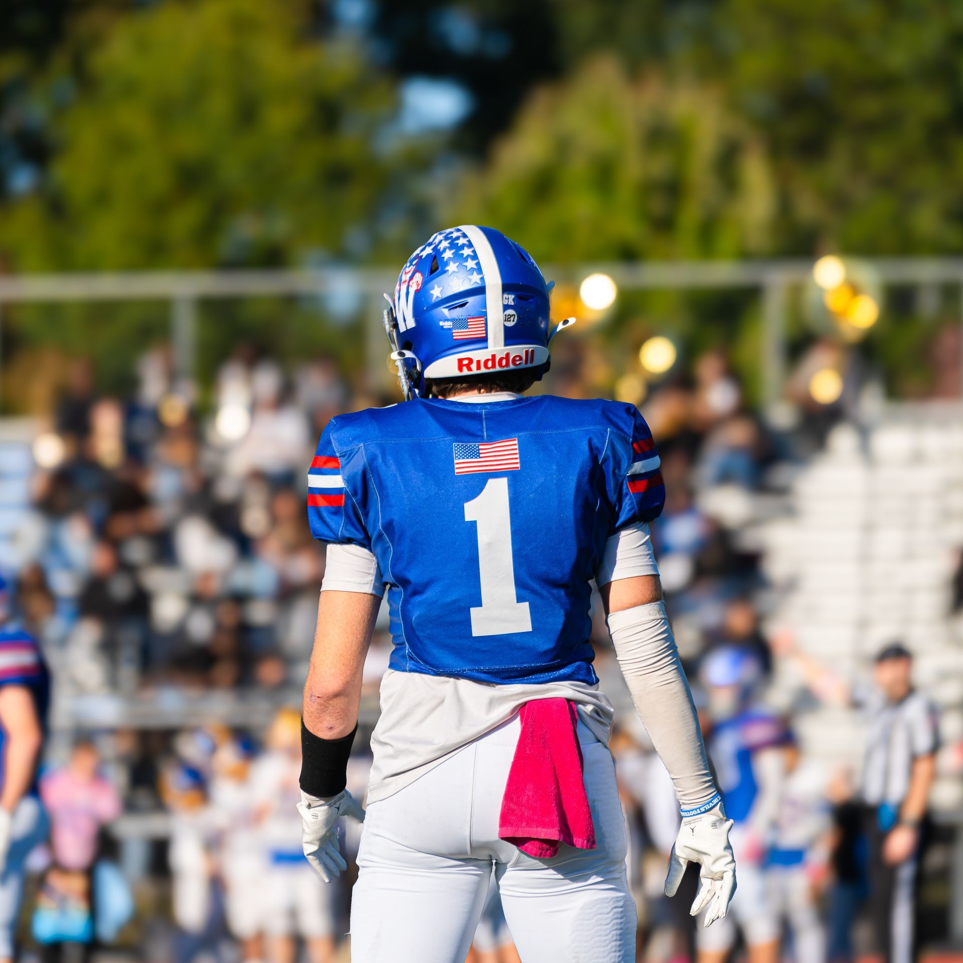 Football player in blue jersey number 1 stands on the field with a pink towel, facing the crowd.