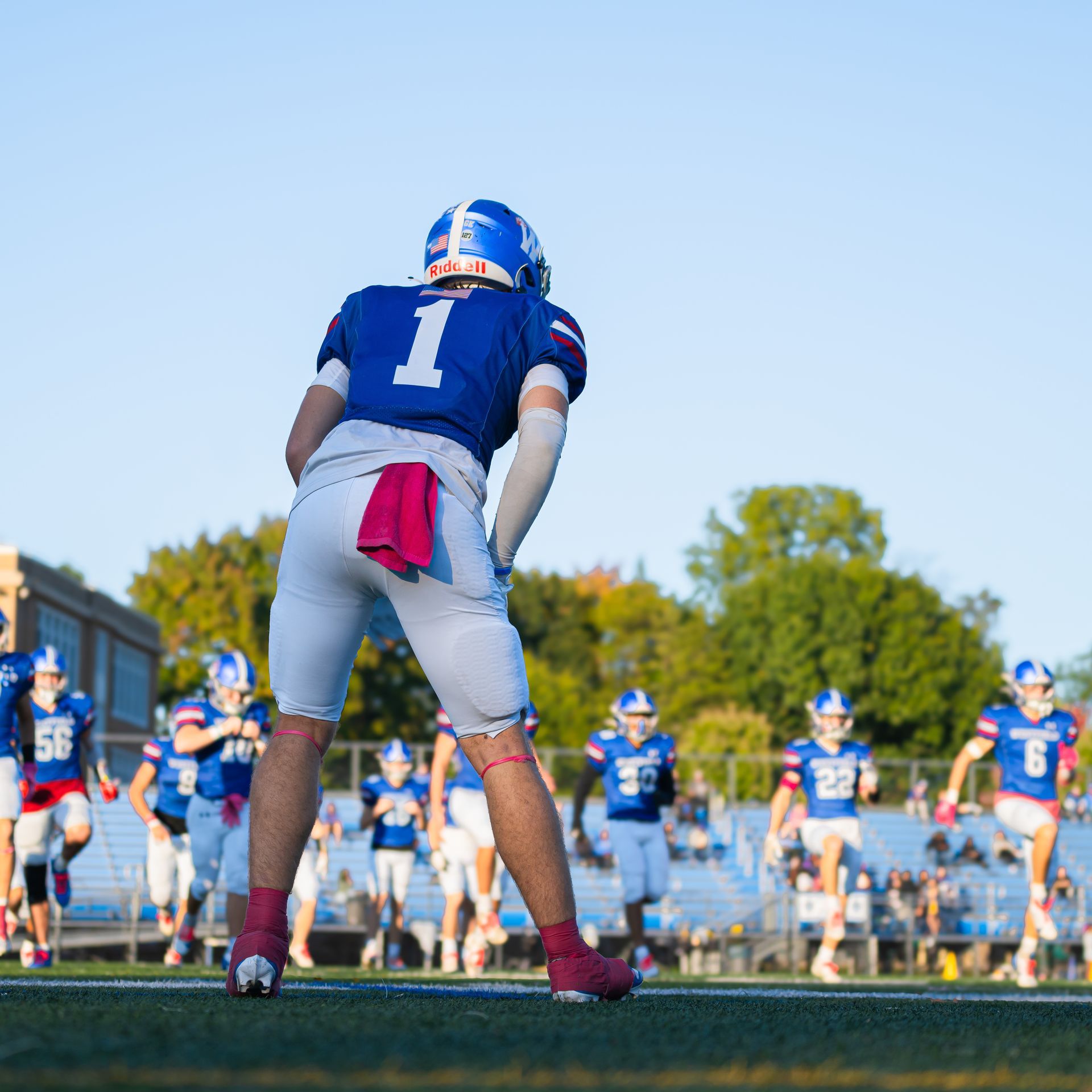 Football player in blue jersey number 1 stands on a field during a game, teammates blurred in the background.