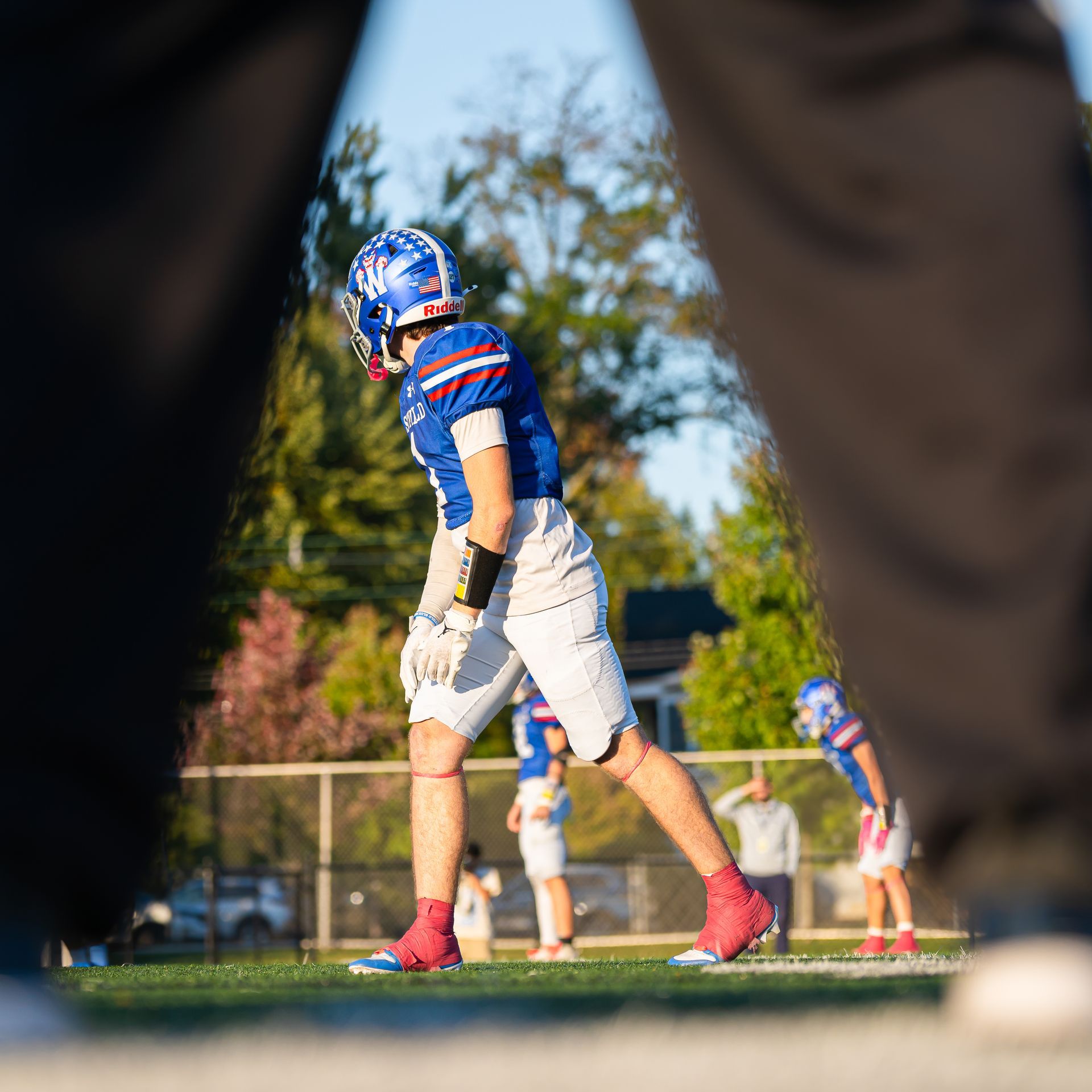 Football player in blue and white uniform walking on field during practice, framed by blurred legs in foreground