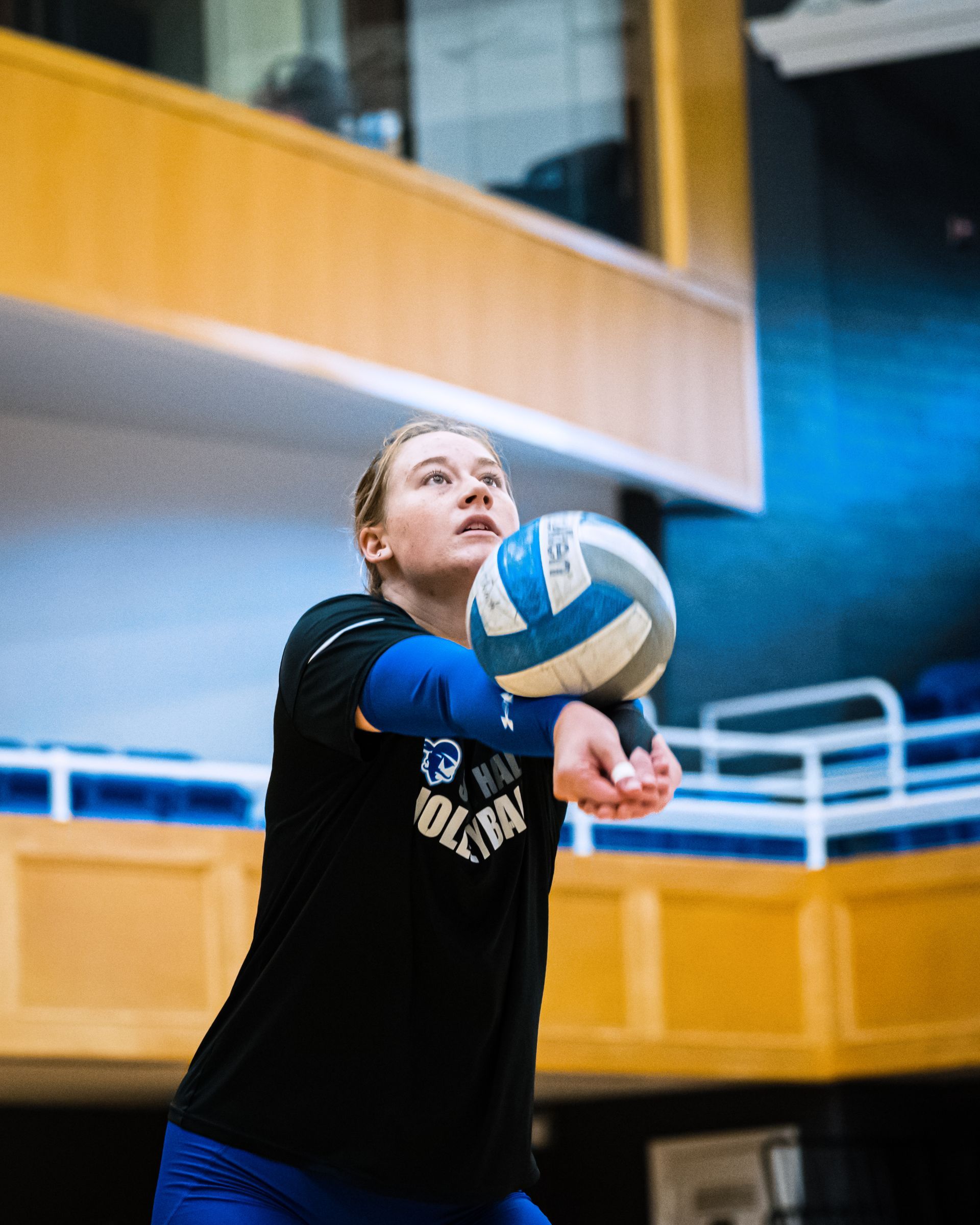 Volleyball player in black shirt and blue arm sleeve forearm-passes a ball indoors