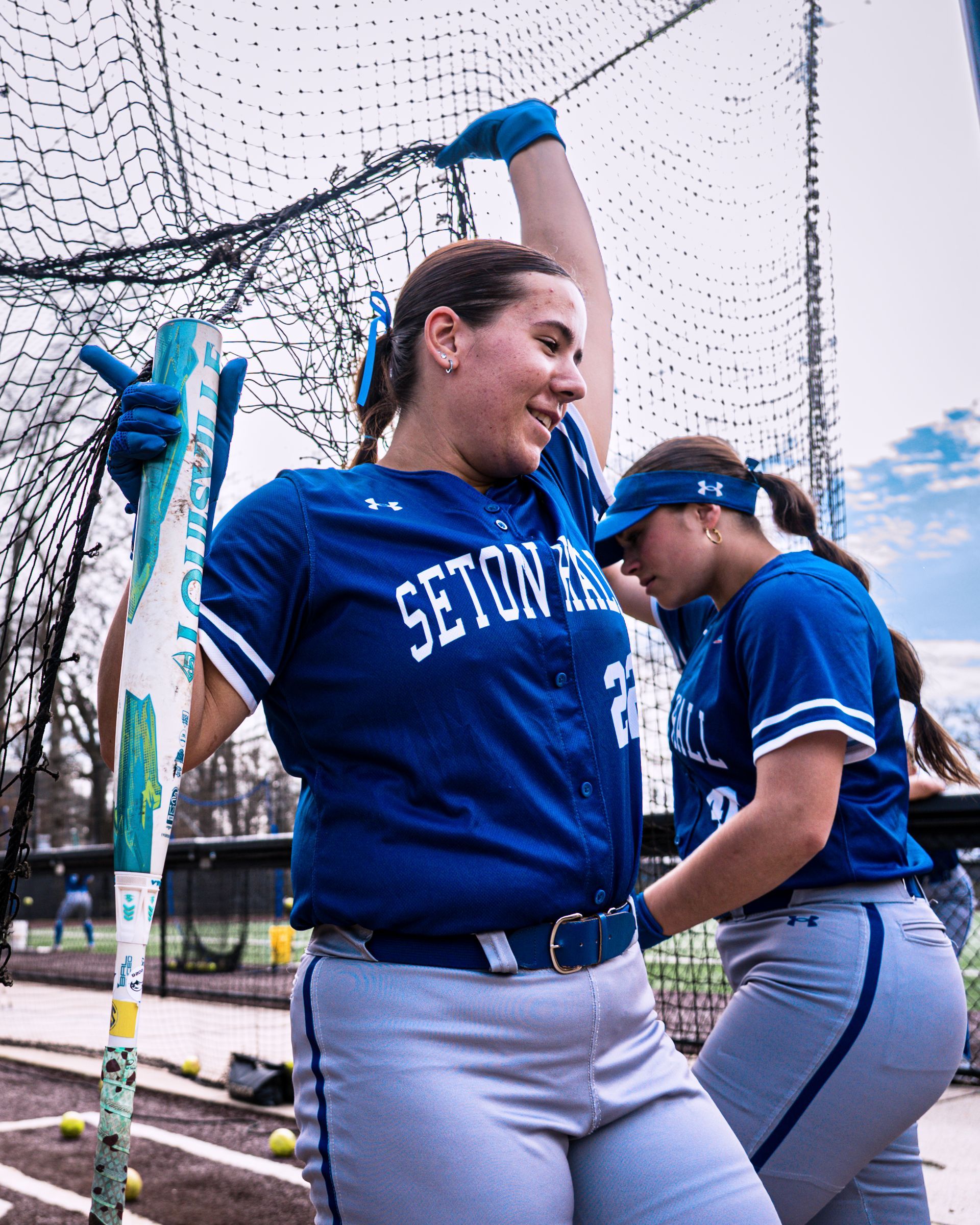 Softball players in blue jerseys stretch beside a net on a field, one raising an arm and smiling.