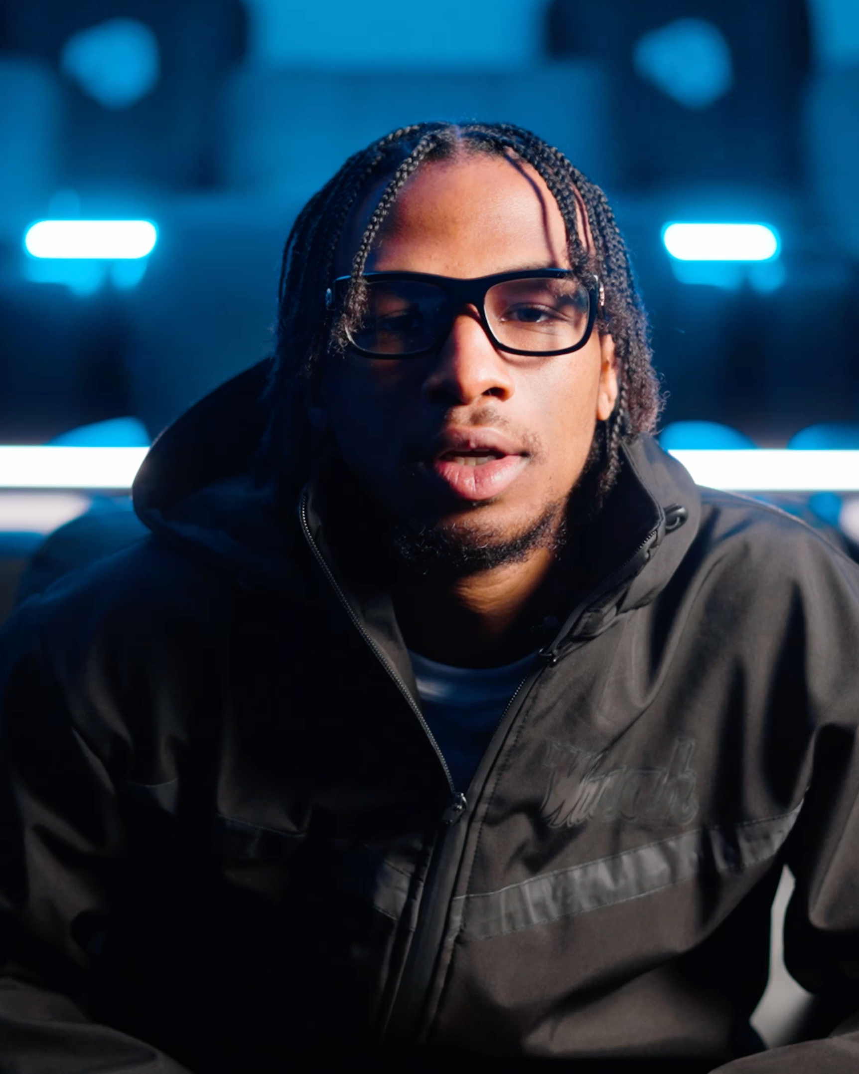 Person with braided hair and glasses in a dark jacket, lit by blue neon lights.