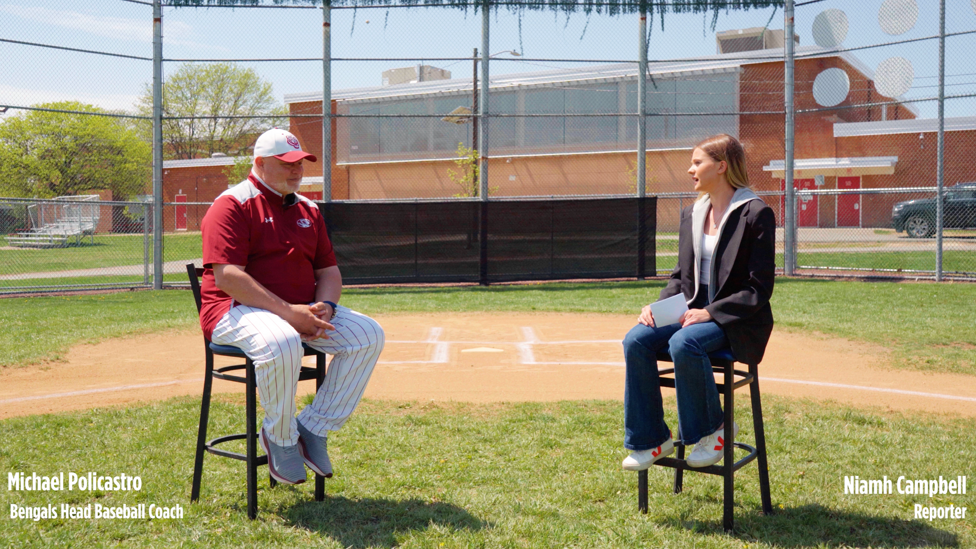 Two people seated on stools talking on a baseball field, with a chain-link backstop and buildings behind them.