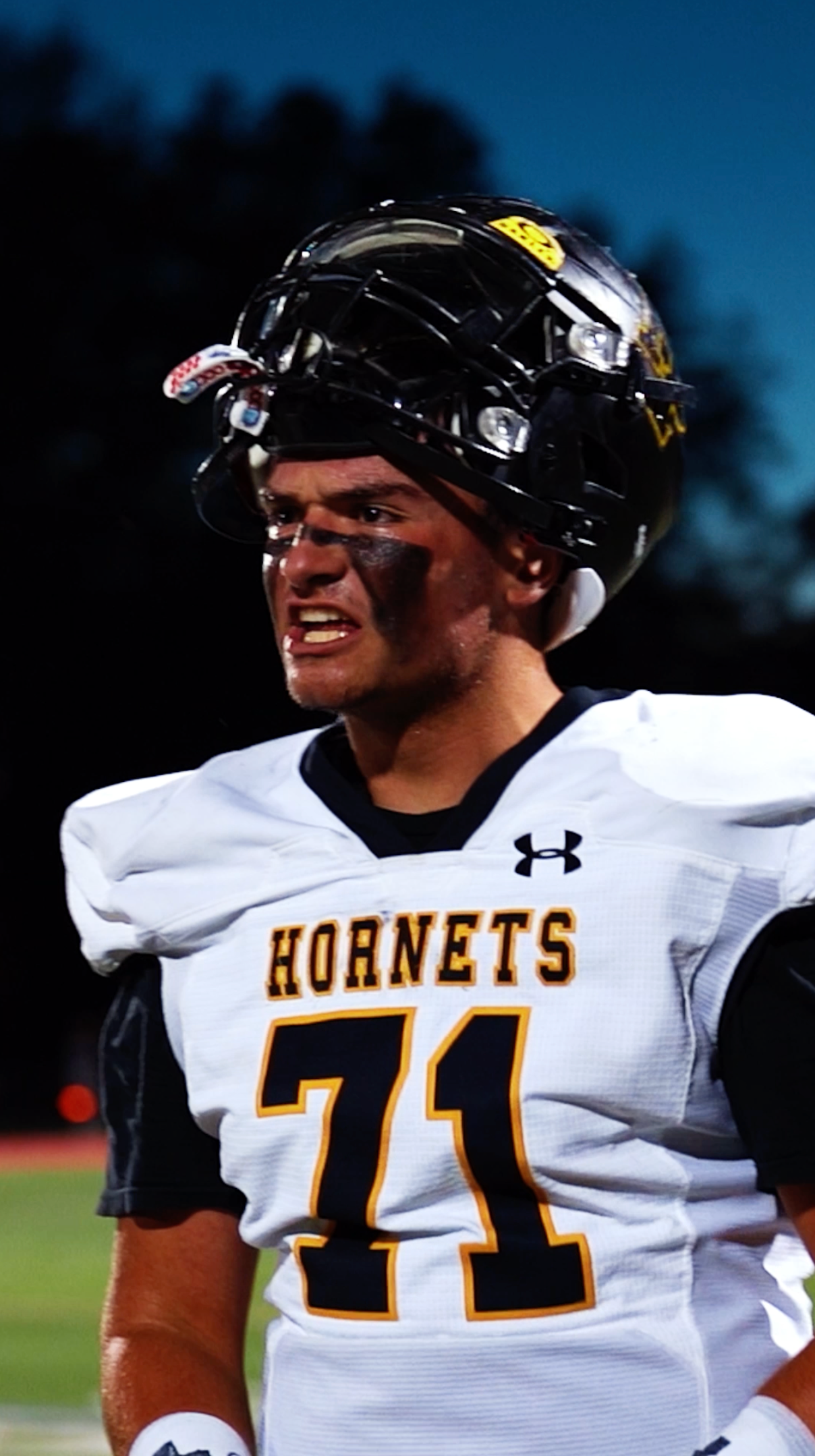 Football player in white Hornets jersey #71 and black helmet, smiling on the field at night