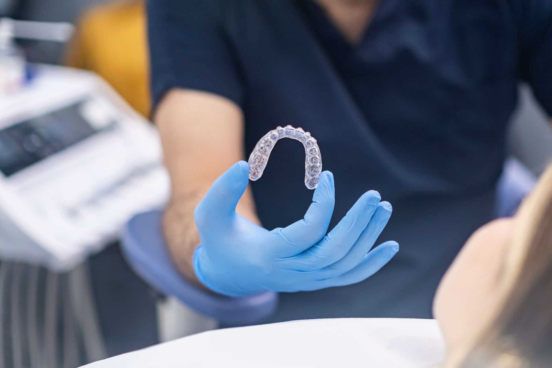 An orthodontist wearing blue rubber gloves and uniform, holding Invisalign in front of a patient. An orthodontist wearing blue rubber gloves and uniform, holding Invisalign in front of a patient.