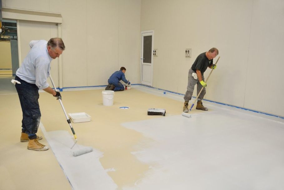 Workers painting a large white room floor with rollers and buckets