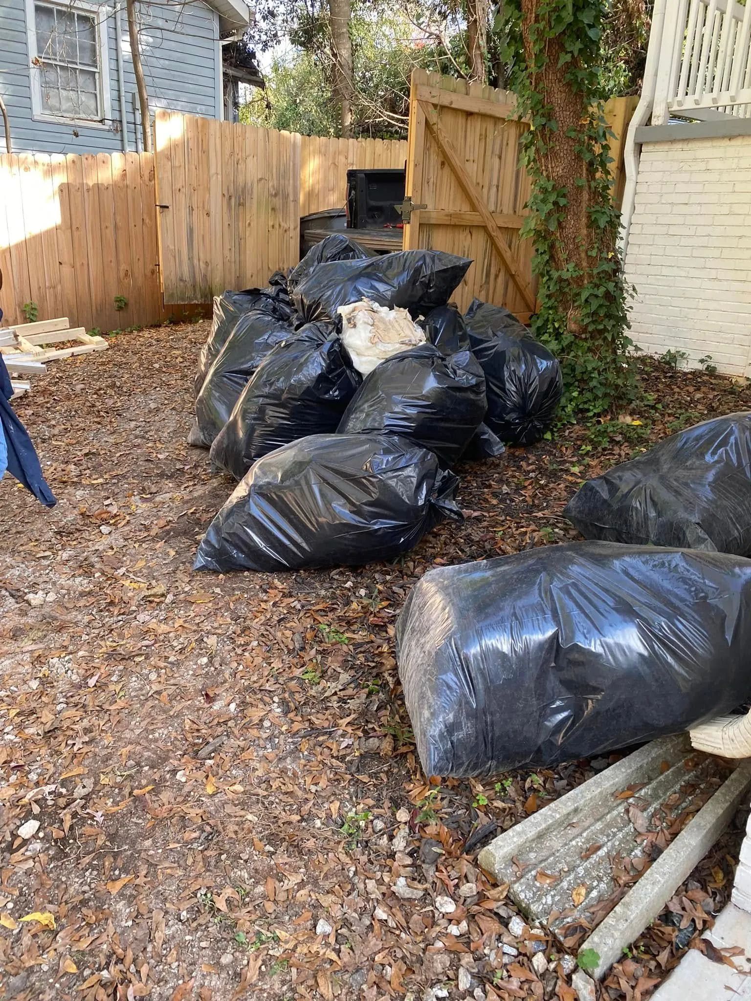 Black trash bags piled in a yard with a wooden fence and tree.