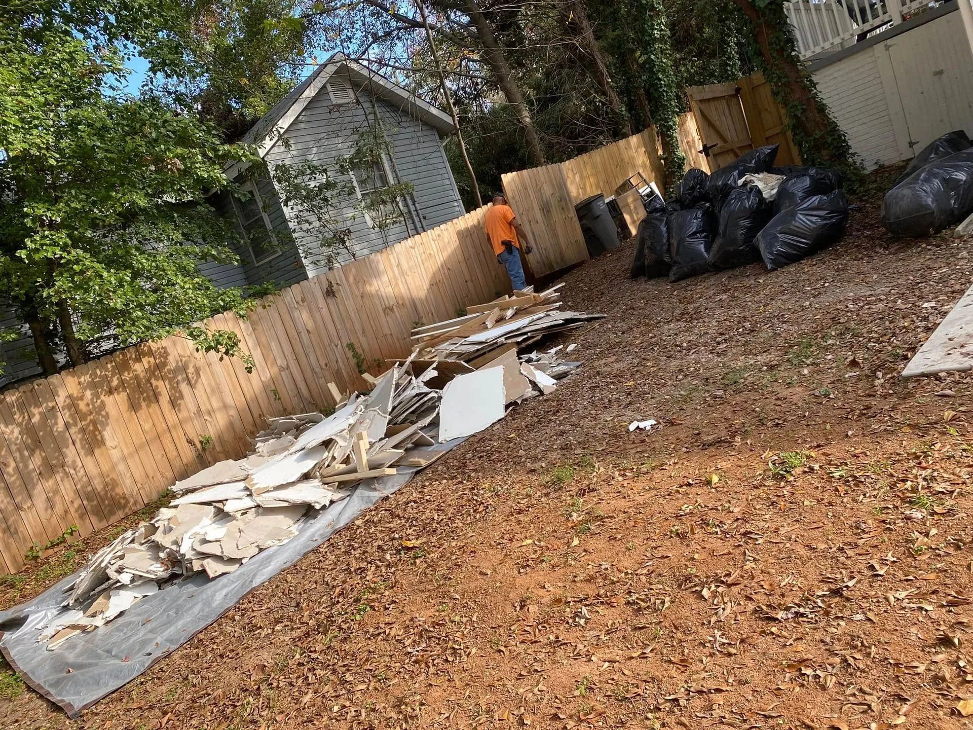 Backyard with debris on ground, worker near fence, black trash bags.