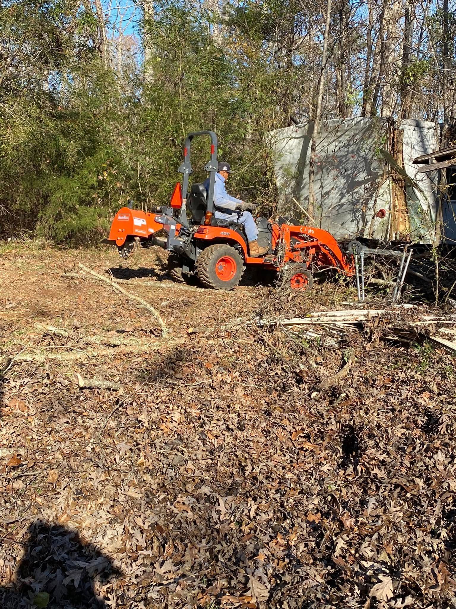 Person operating an orange tractor in a wooded area, clearing debris with a front-end loader.