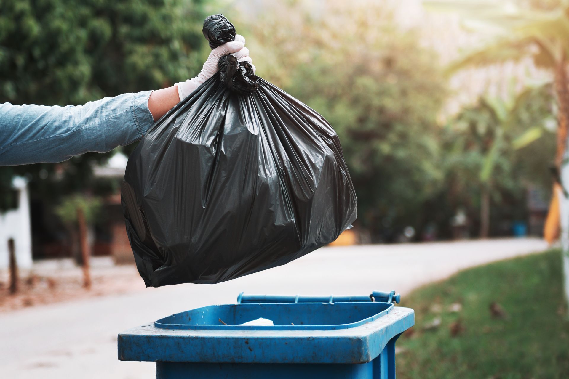 Person in glove throws a black garbage bag into a blue trash can outdoors, near trees.