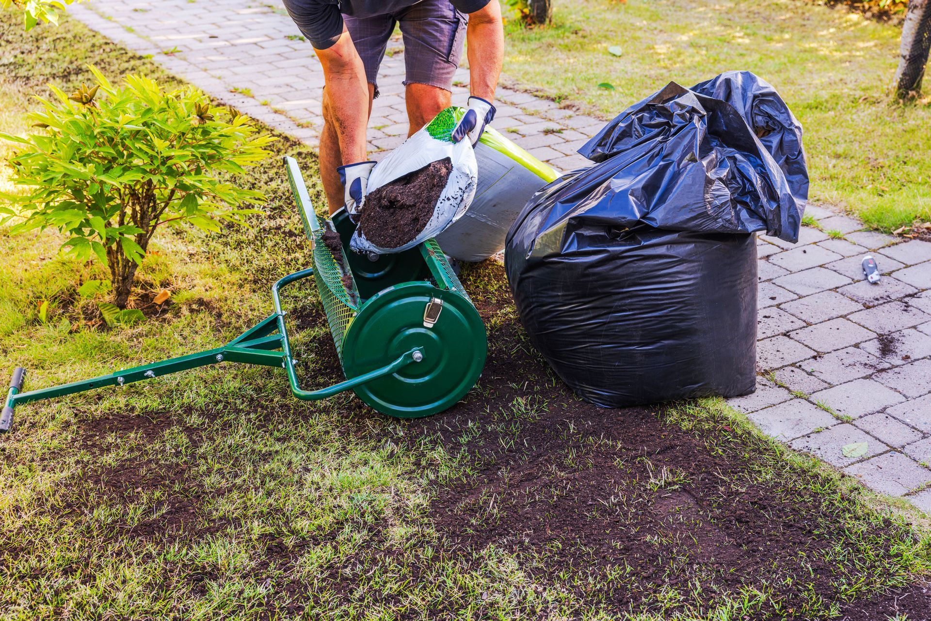 Man spreading soil, fertilizer, or seed near a small lawn roller, black trash bag, and a sidewalk.