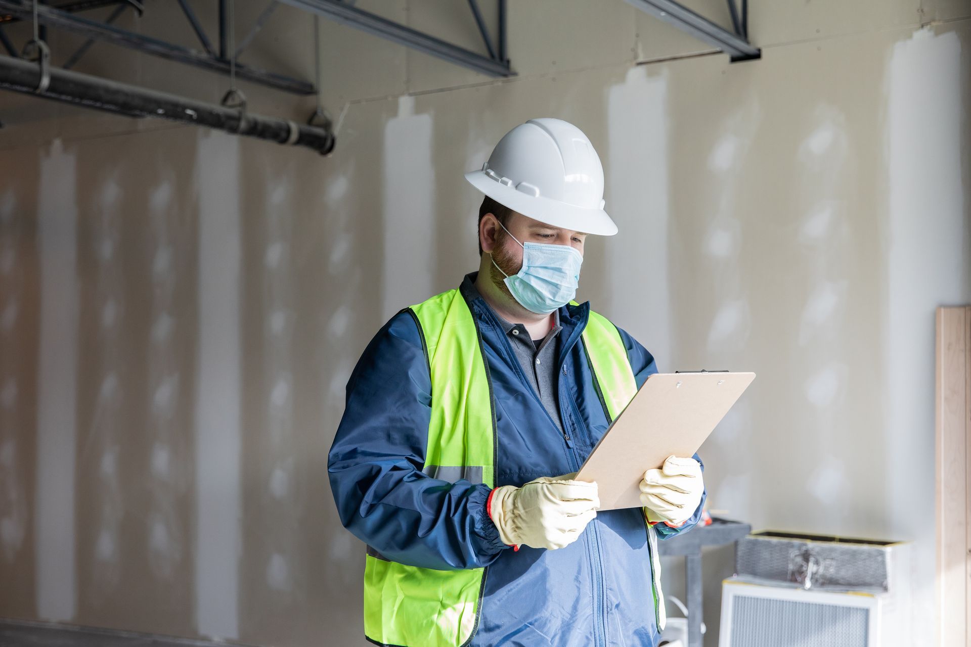 A Construction Worker Wearing a Mask and Hard Hat is Looking at a Clipboard - Fire Sprinkler Services in Fort Wayne, IN