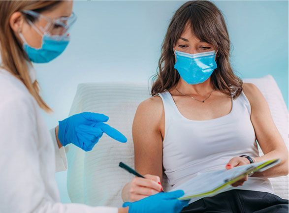 A woman wearing a mask is sitting in a hospital bed while a doctor writes on a clipboard.