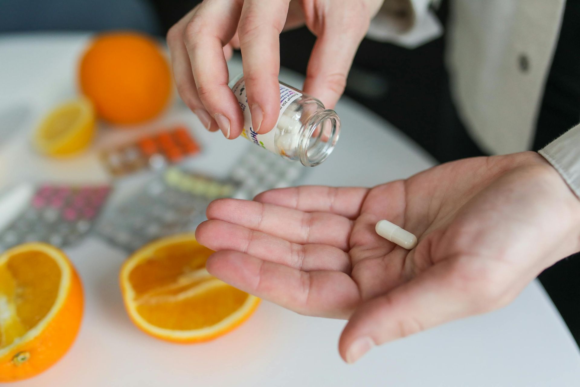 Person pouring a capsule from a bottle into their hand. Orange slices and pills are in the background.