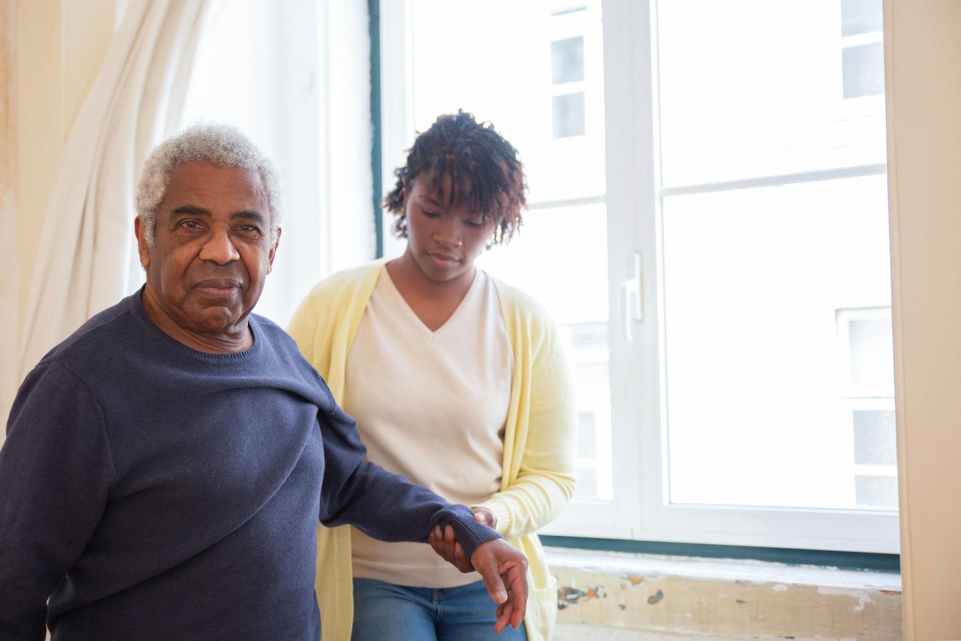 A woman is helping an elderly man walk in front of a window.
