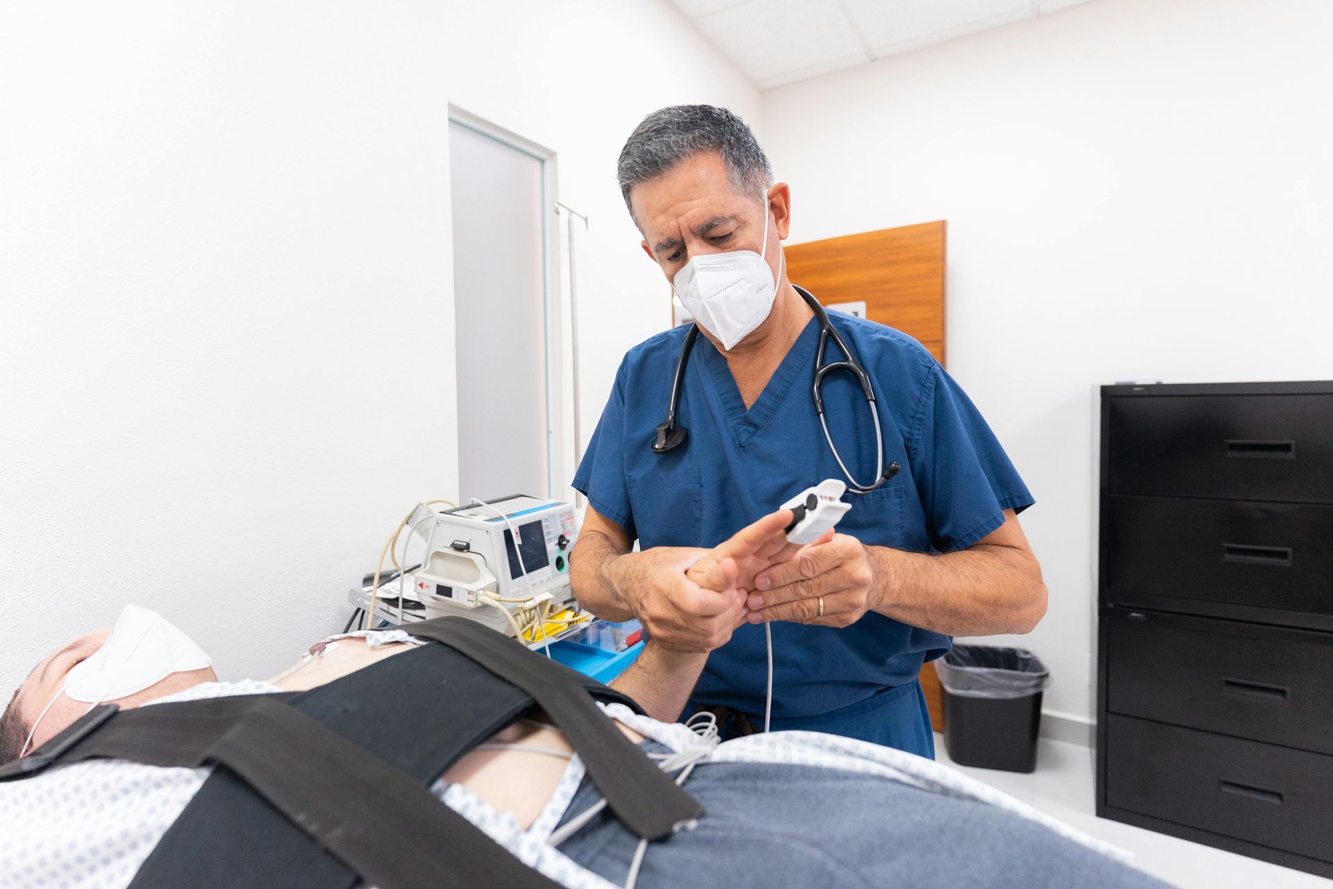 A doctor wearing a mask is examining a patient 's arm in a hospital room.