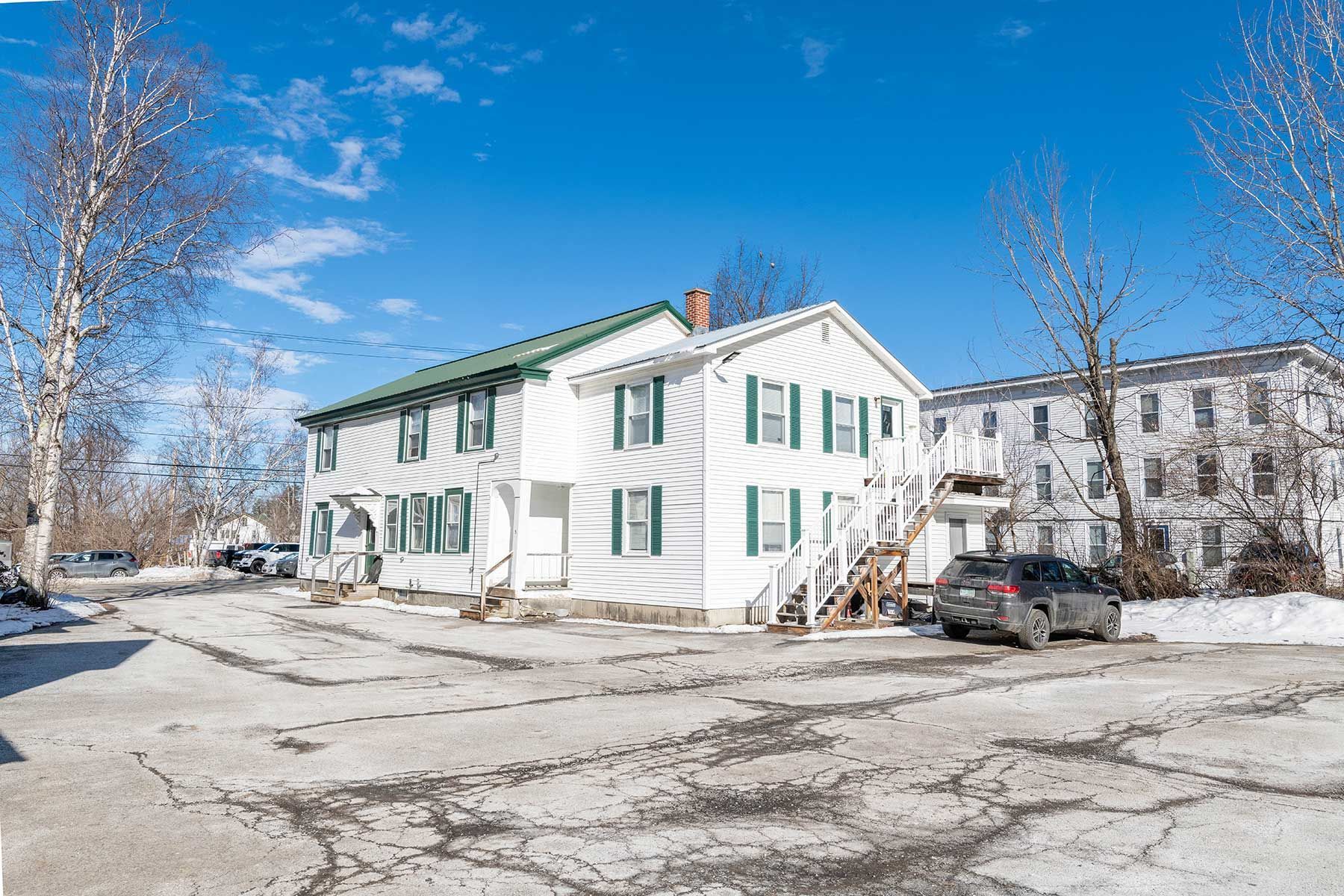 White two-story house with green trim and parked cars in a snowy lot at Elm Complex on a clear day.