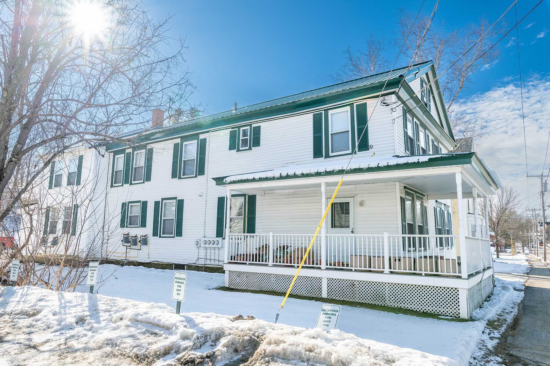 Snow-covered white house with green shutters and porch under a bright blue winter sky at Elm Complex in New Hampshire.