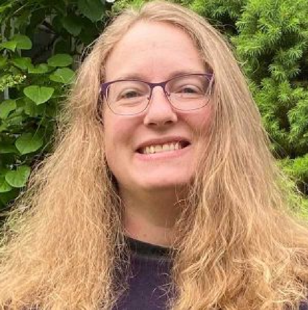 Smiling person with long blond hair and glasses, standing outdoors in front of green foliage