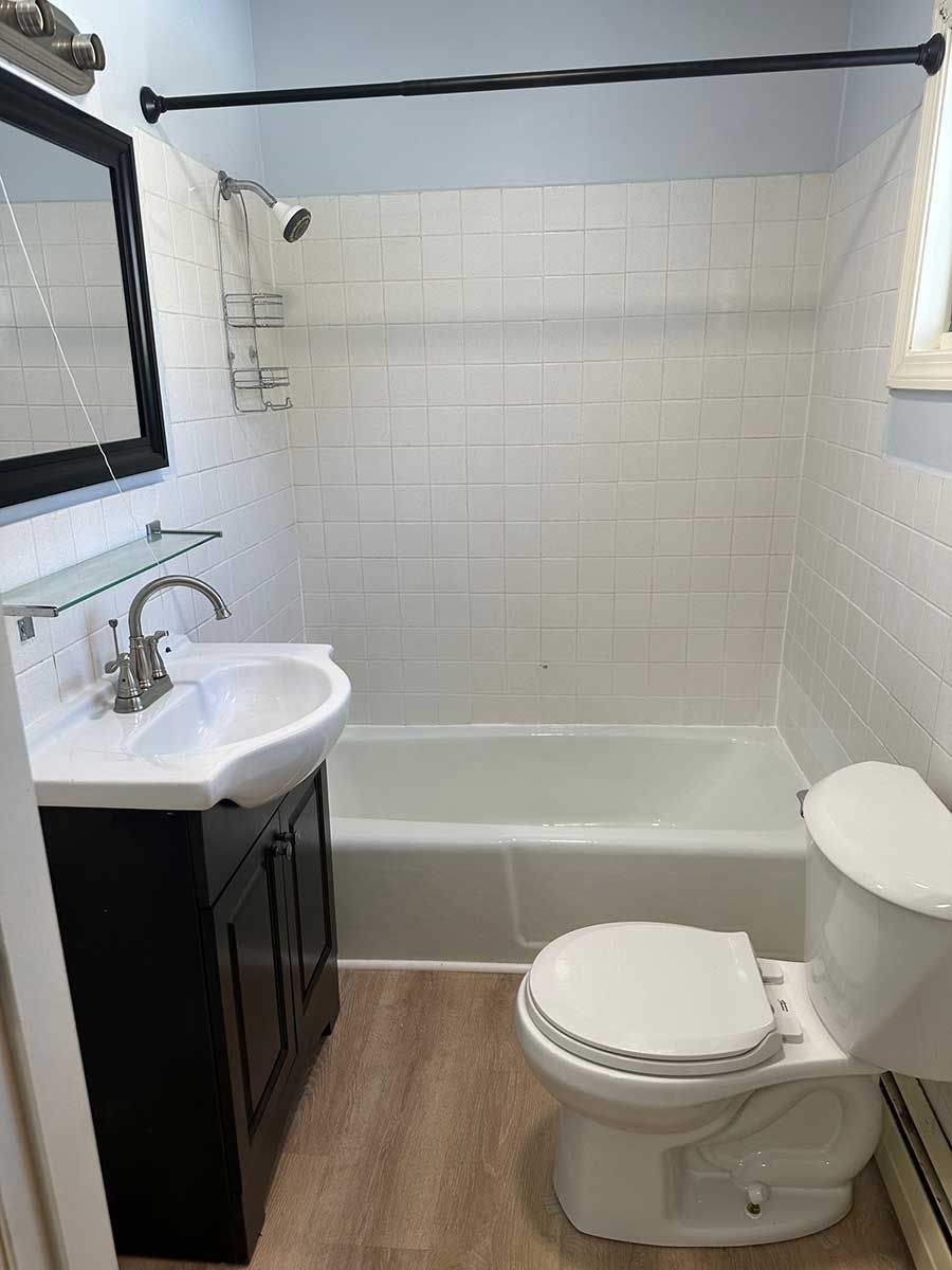Small white bathroom at Bickford Hill Complex with pedestal sink, toilet, tiled shower wall, and wood-look flooring.