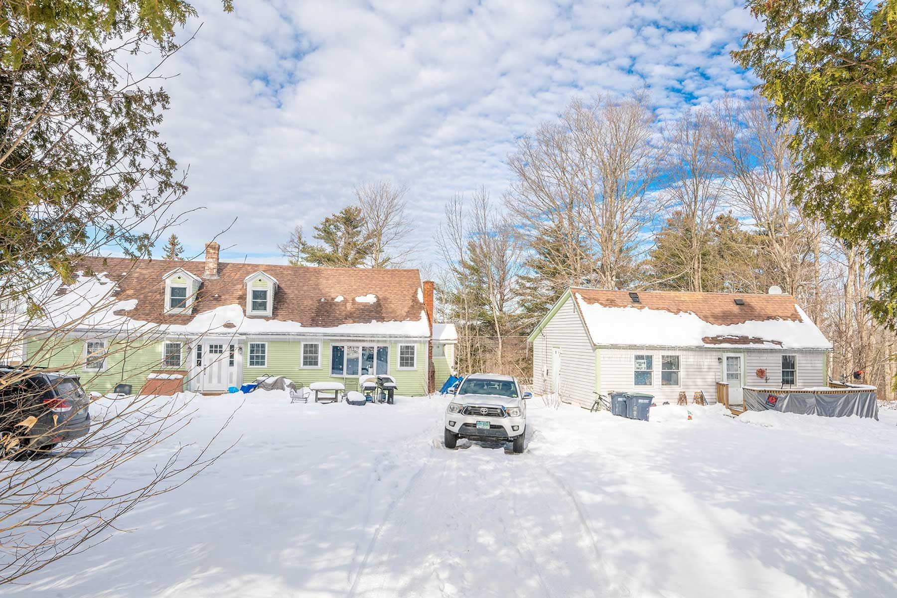Snow-covered house and driveway with parked truck, leafless trees under a blue sky