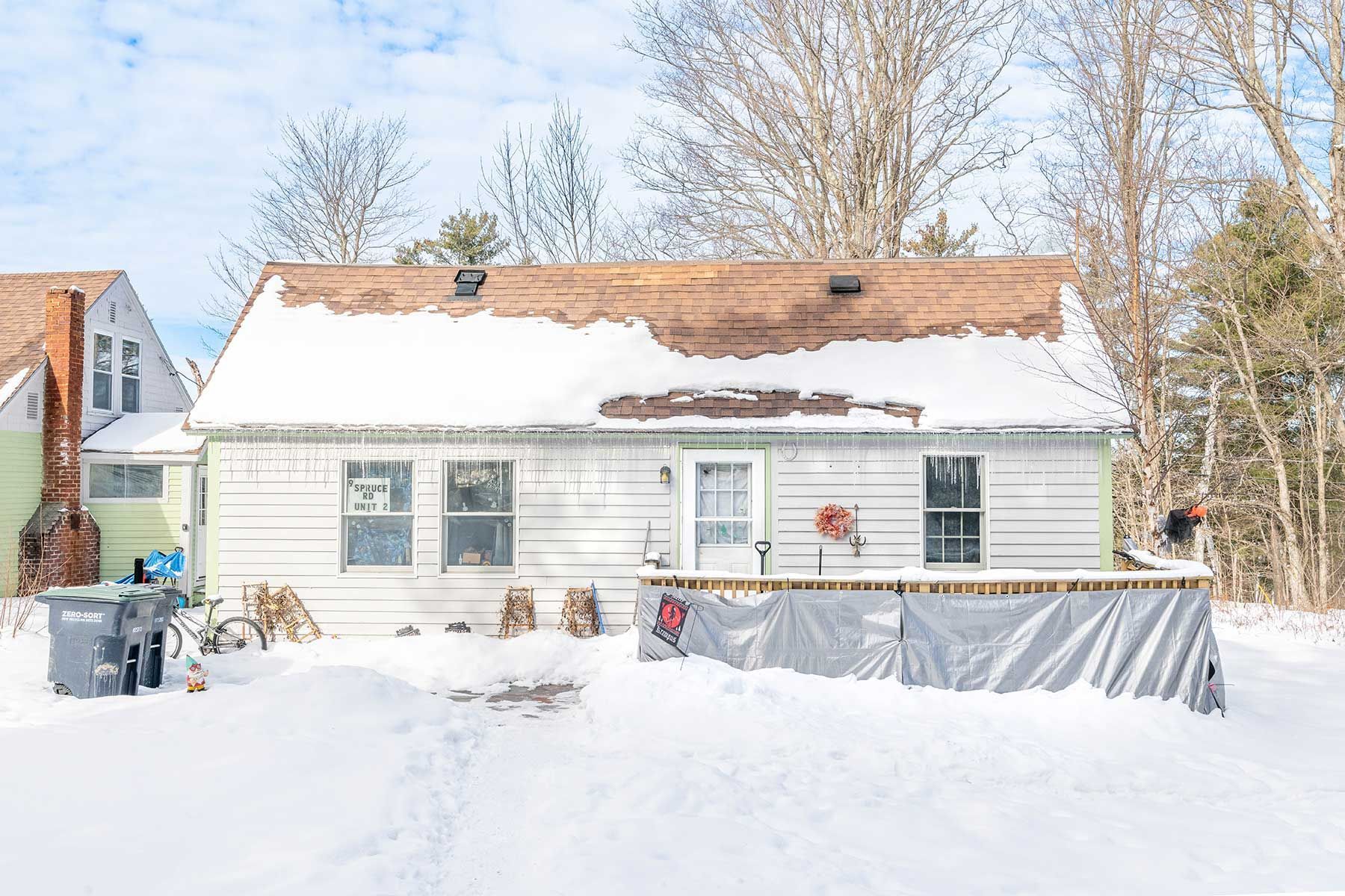 Snow-covered white house with partially snow-covered roof and fenced backyard in winter