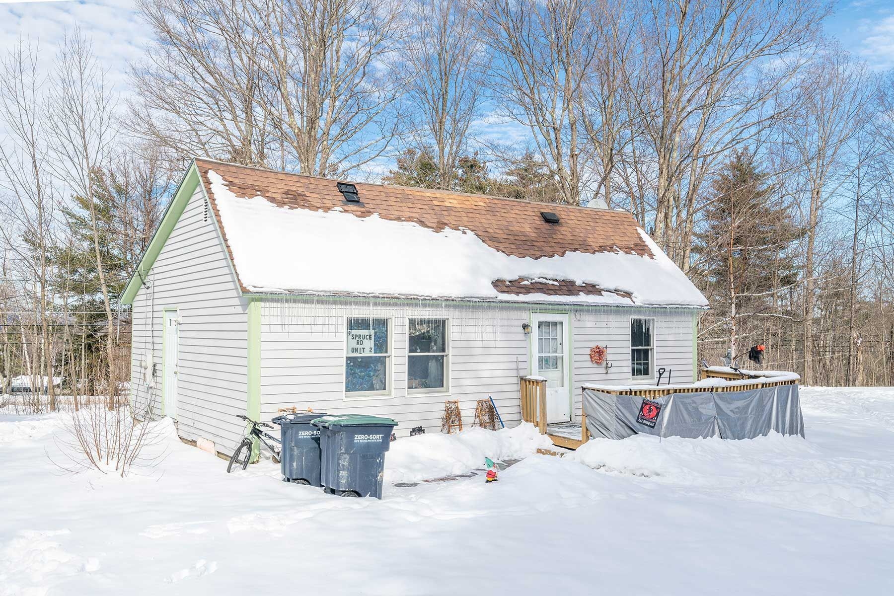 Snow-covered white house with damaged roof and blue trash bins in winter yard